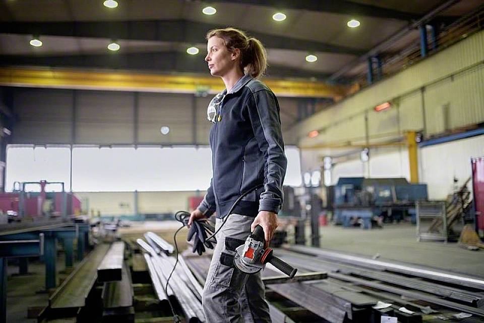 Een vrouw in werkkleding staat in een grote industriële fabriekshall en houdt een elektrisch gereedschap vast.