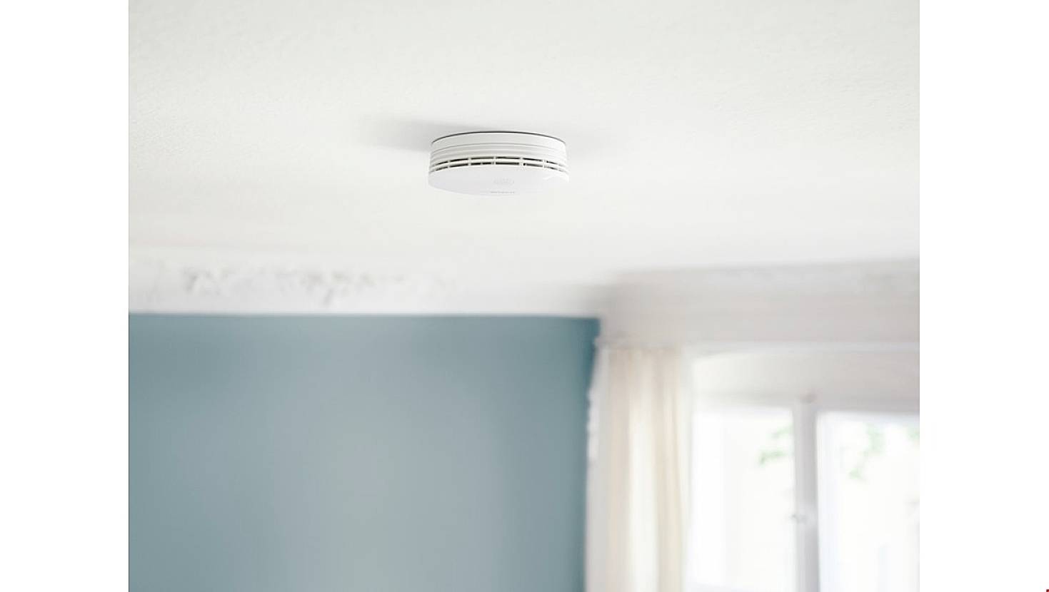 Een rookmelder is aan het plafond bevestigd in een kamer met een blauwe muur en een raam.