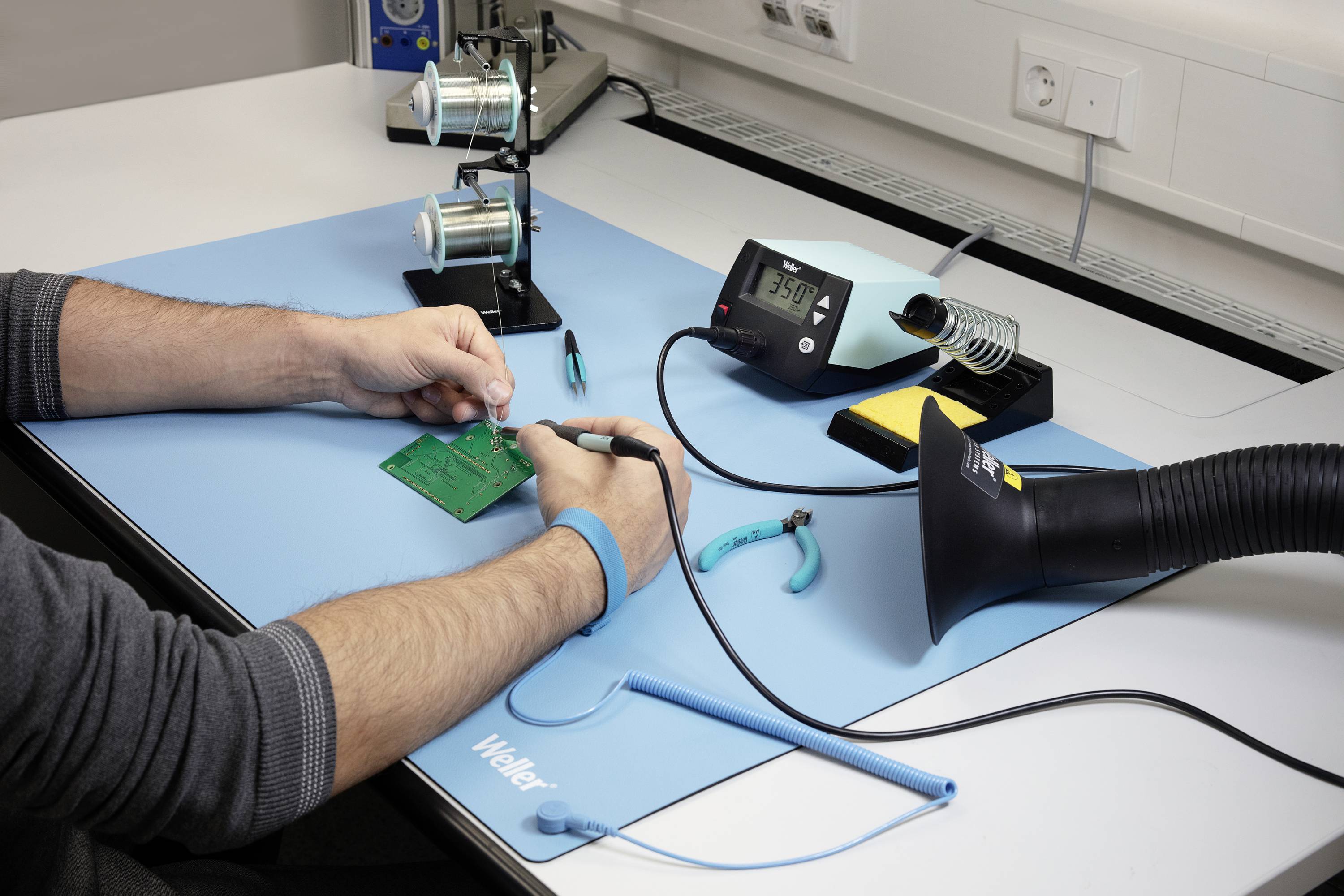 Een persoon soldeert een groene printplaat op een blauwe tafel met een soldeerbout. Een antistatische polsband, ventilator en gereedschappen zijn eveneens zichtbaar.