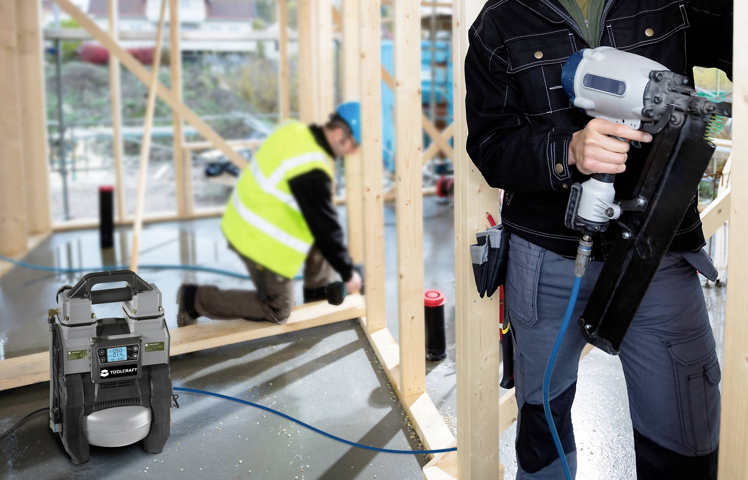 Een persoon werkt op een bouwplaats met een pneumatische spijkerpistool. Op de voorgrond staat een draagbare compressor. Een arbeider op de achtergrond.