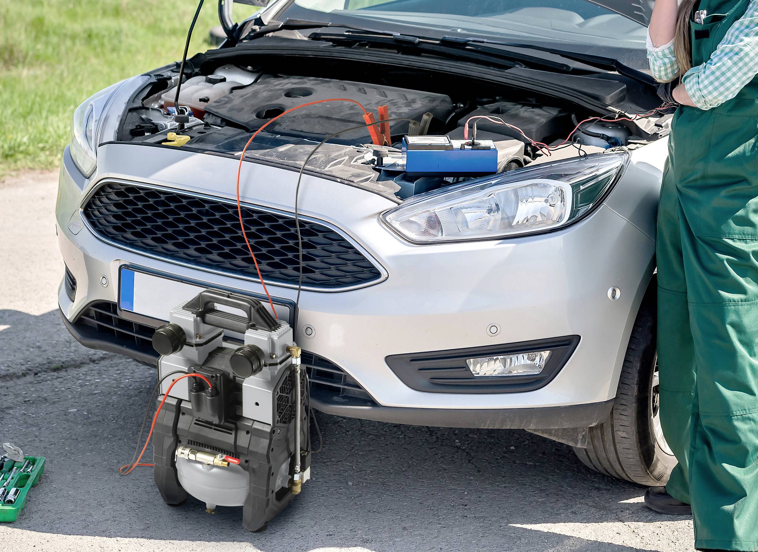Een monteur staat naast een auto met de motorkap open. Startkabels zijn aangesloten op de autobatterij, er wordt een hulpstart gegeven.