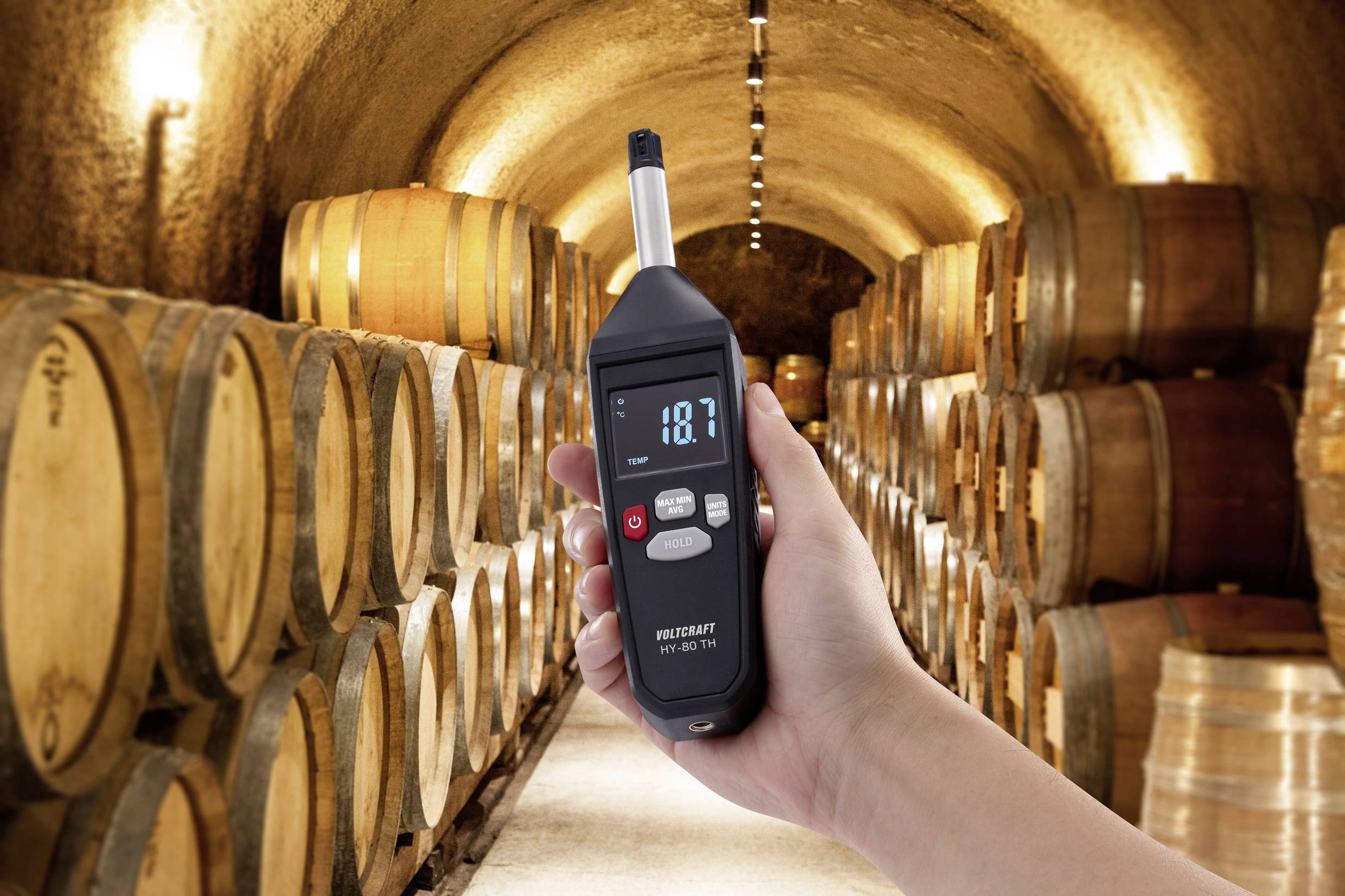 Een hand houdt een digitale vochtigheidsmeter vast met de aanduiding '18.7' in een wijnkelder vol houten vaten.
