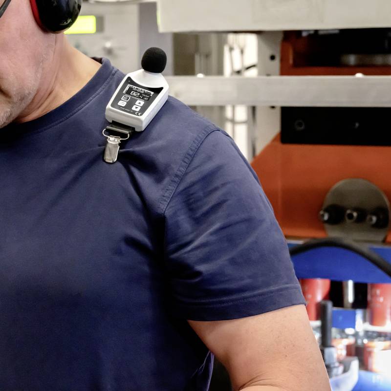 Een man draagt een microfoon en een draagbaar apparaat, mogelijk voor communicatie of geluidsopname, in een industriële omgeving.