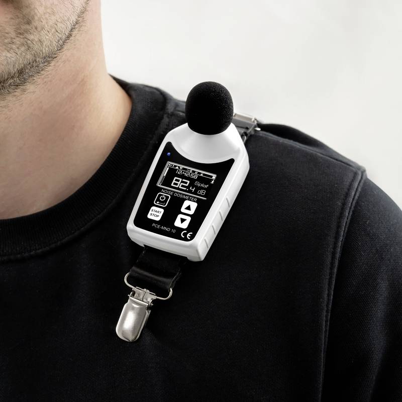 Een man draagt een decibel-meetapparaat aan zijn trui. Het display toont 82,4 dB met een batterijstatus en microfoon-pictogram.