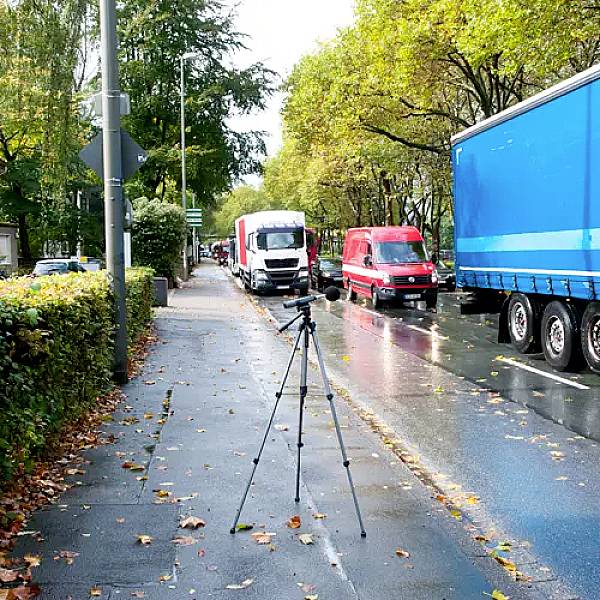 Een statief staat op een natte stoep naast een weg. Verschillende geparkeerde bestelwagens en een vrachtwagen zijn te zien op de straat.