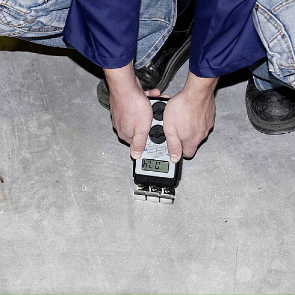 Een persoon in spijkerbroek en veiligheidsschoenen houdt een krachtmeetapparaat vast dat een waarde aangeeft op een betonnen vloer.