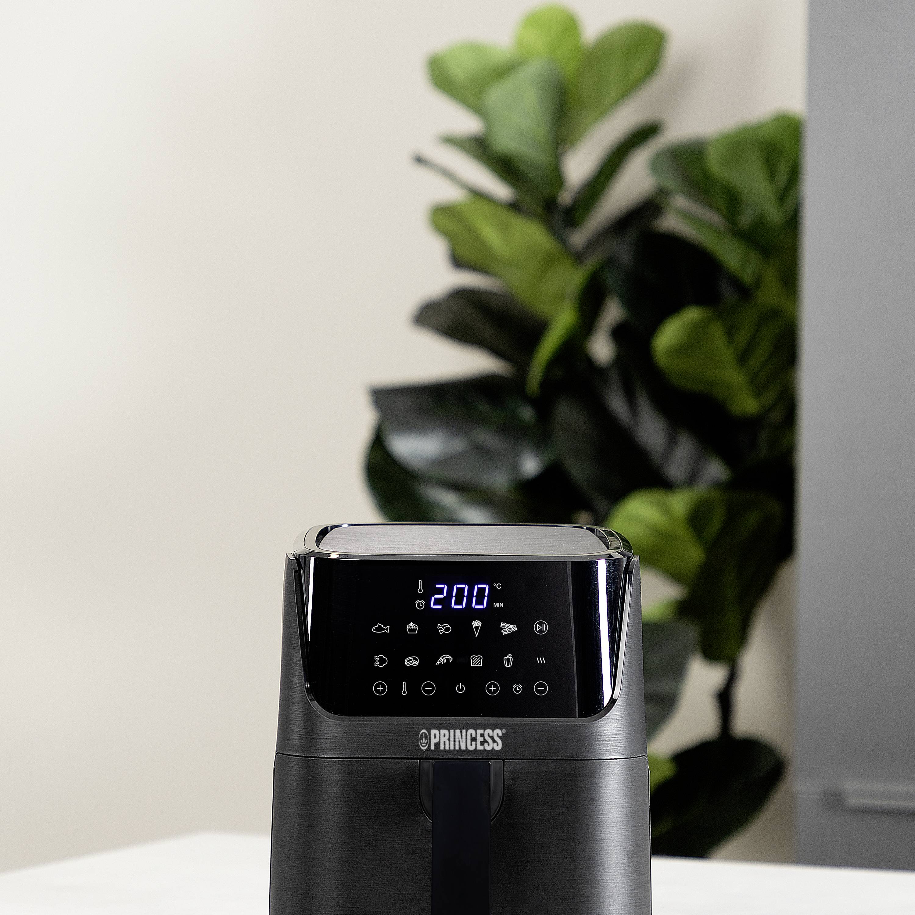 Een zwarte airfryer met een digitaal display dat verschillende symbolen toont. Op de achtergrond zijn groene planten zichtbaar.