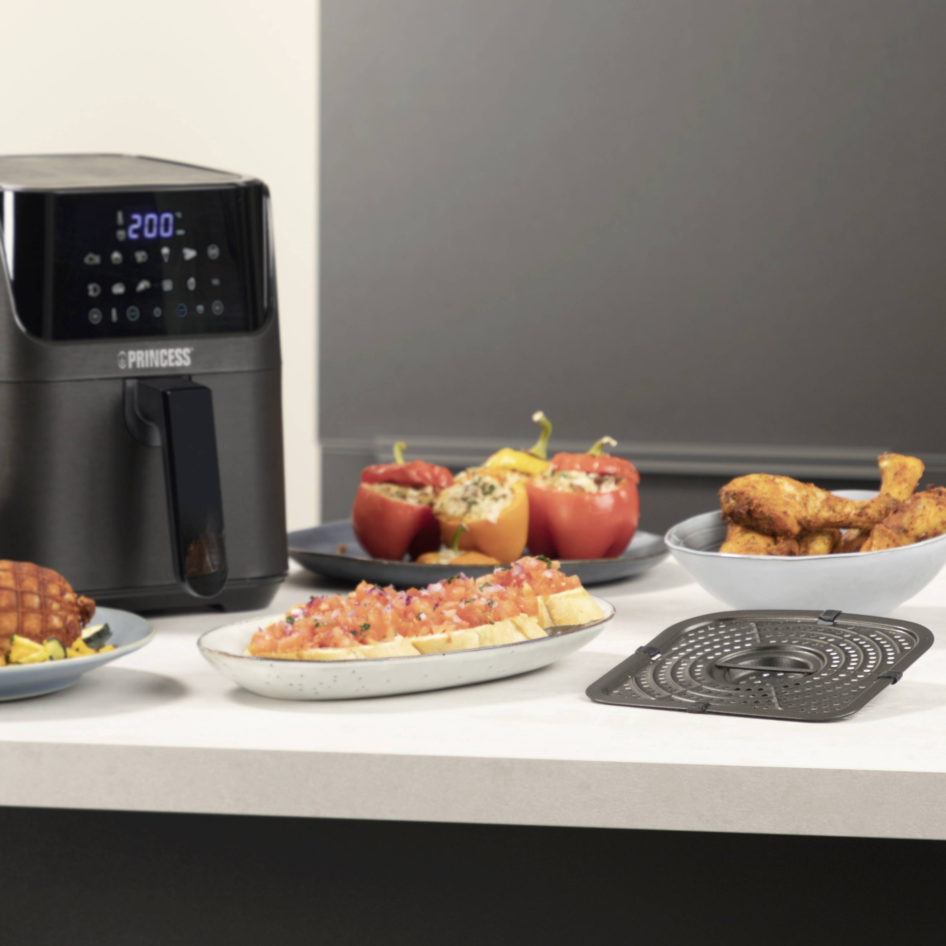 Een heteluchtfriteuse op een aanrecht, omgeven door gevulde paprika's, friet, een stuk wafel en een tomatengerecht.