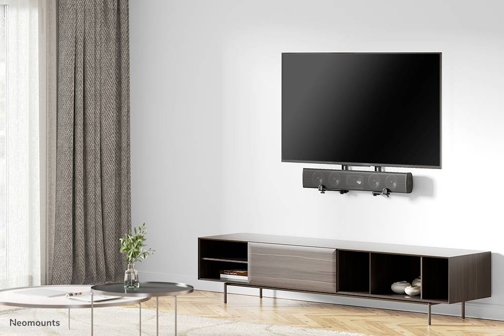 Een woonkamer met een flatscreen-tv aan de muur, bevestigd boven een zwarte luidspreker, naast een houten tafel en een dressoir.