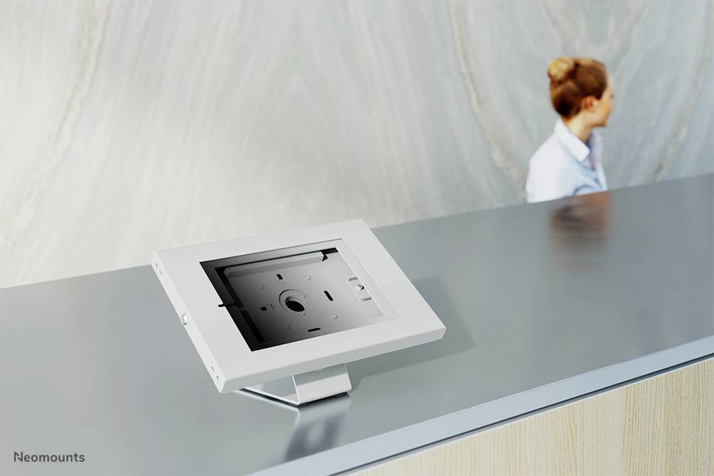 Een vrouw staat achter een grijze balie waarop een zilveren tablethouder zonder tablet staat. De omgeving oogt modern.