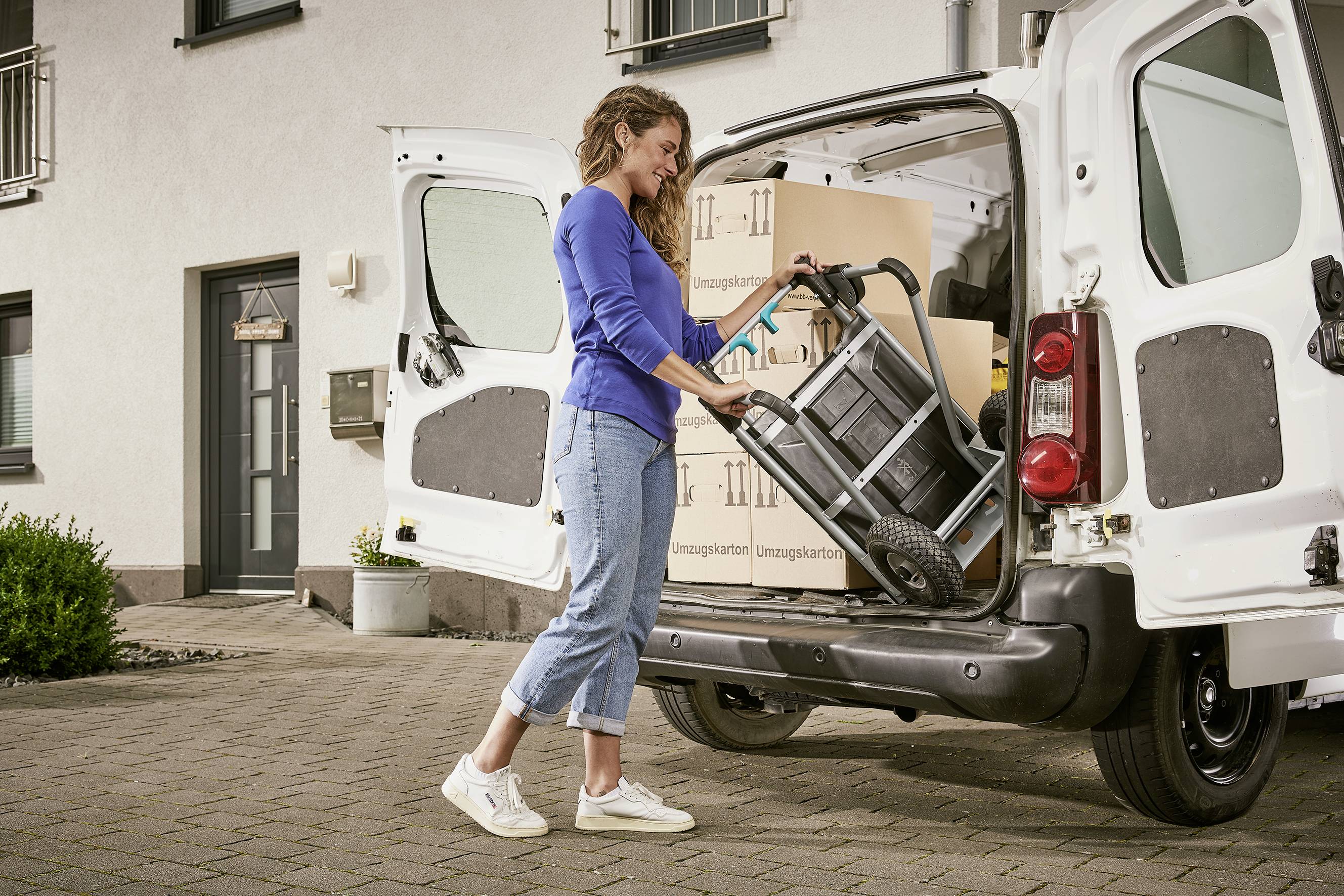 Een vrouw lost dozen uit een bestelwagen met een steekwagen voor een huis. De scène toont een verhuizing of levering.