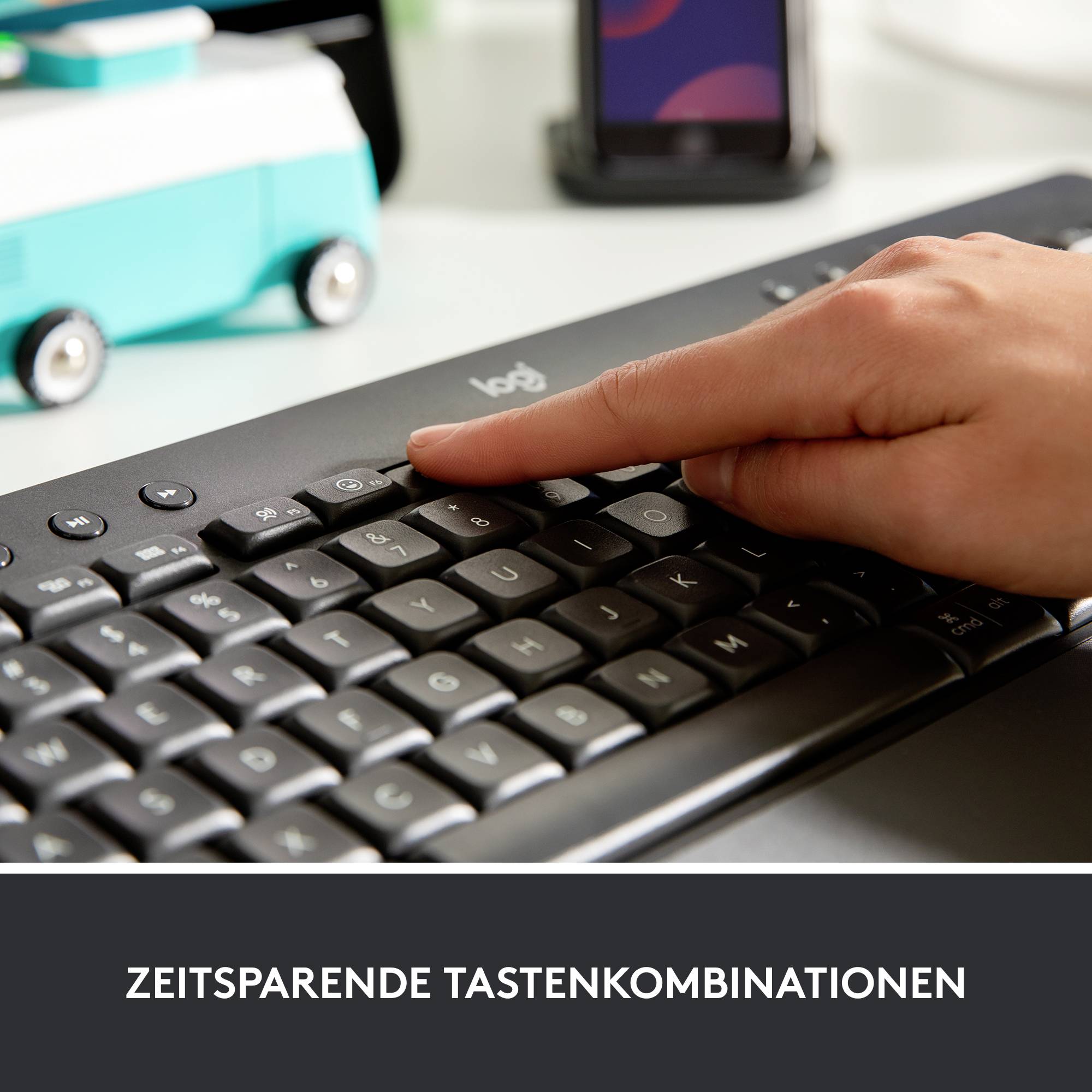 Een hand typt op een computertoetsenbord. Op de achtergrond is een onscherpe smartphone zichtbaar.