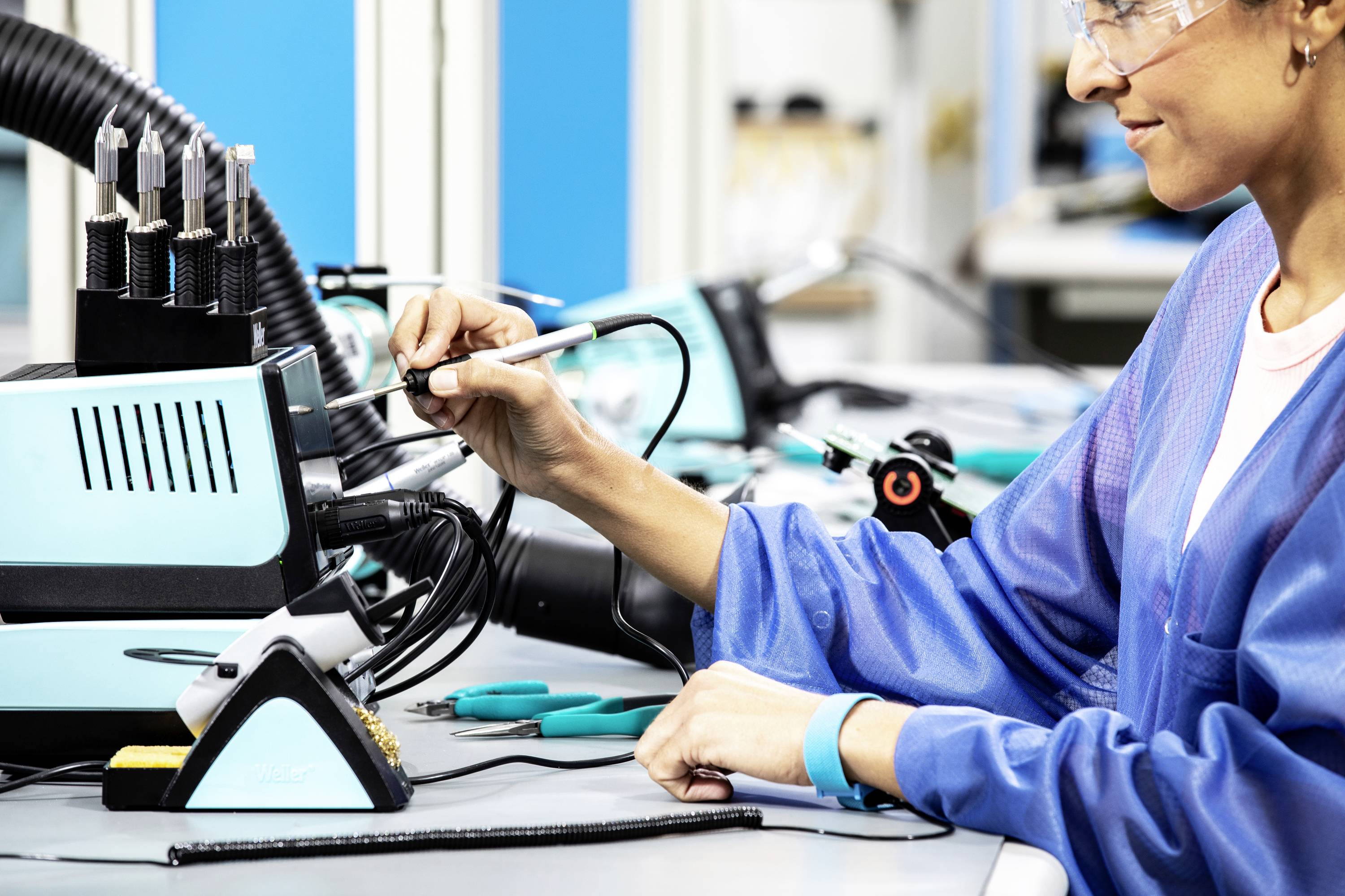 Een persoon in blauwe beschermende kleding lost kabels bij een soldeerpunt met een soldeerbout in een elektronicawerkplaats.
