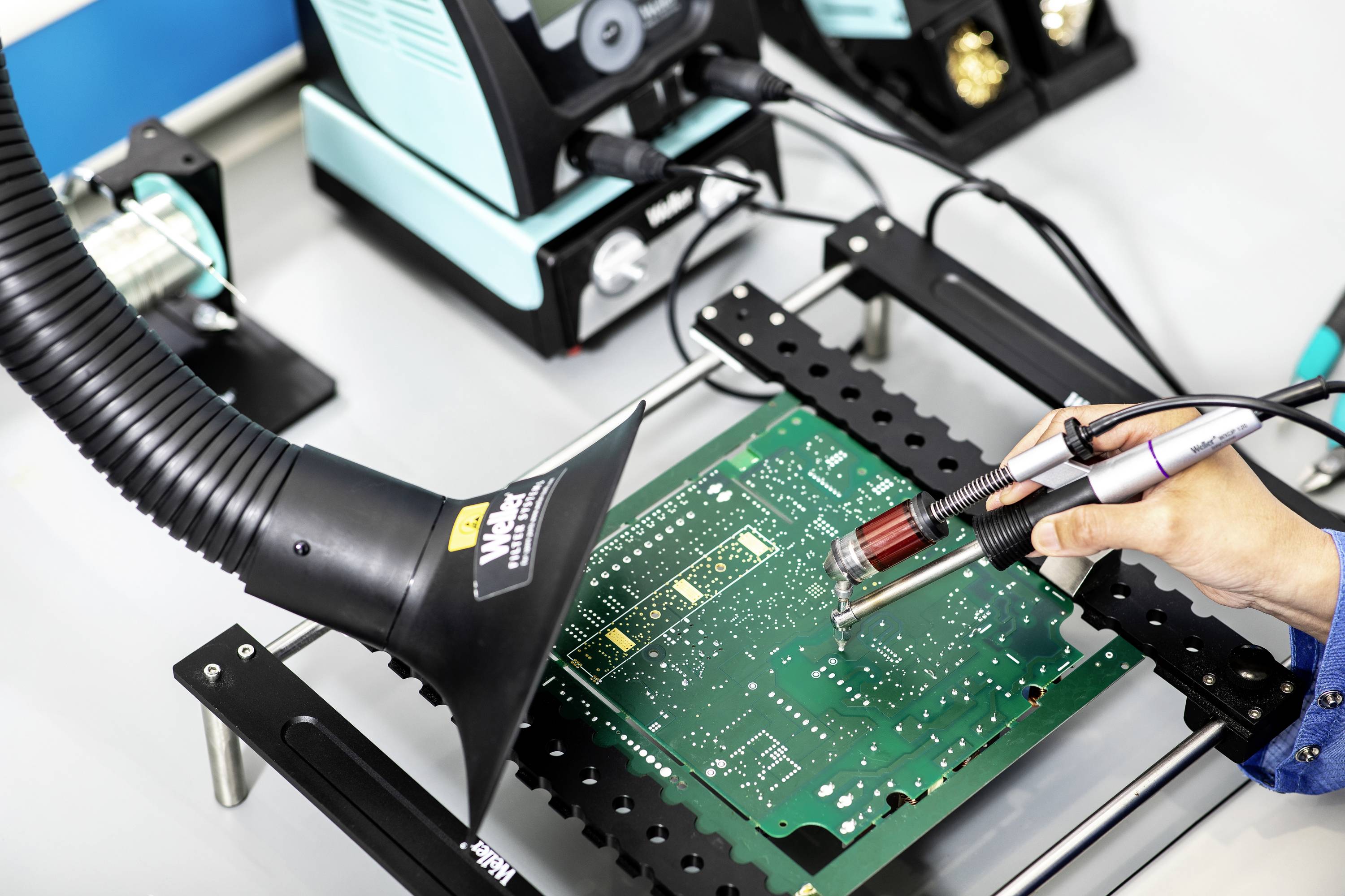 Een persoon soldeert een printplaat, waarbij een afzuigarm zich in de nabijheid bevindt. Op de achtergrond is soldeeruitrusting te zien.
