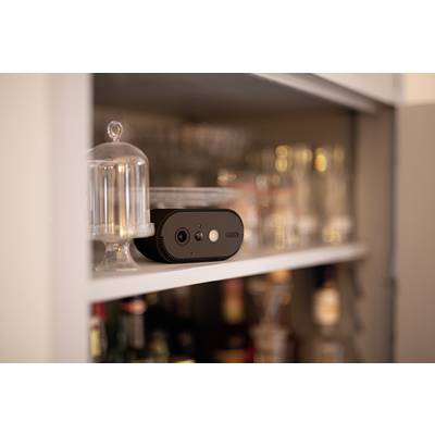 Een kleine, zwarte camera staat op een plank in een keuken, omringd door glazen. Hij wordt gebruikt voor bewaking of opname.