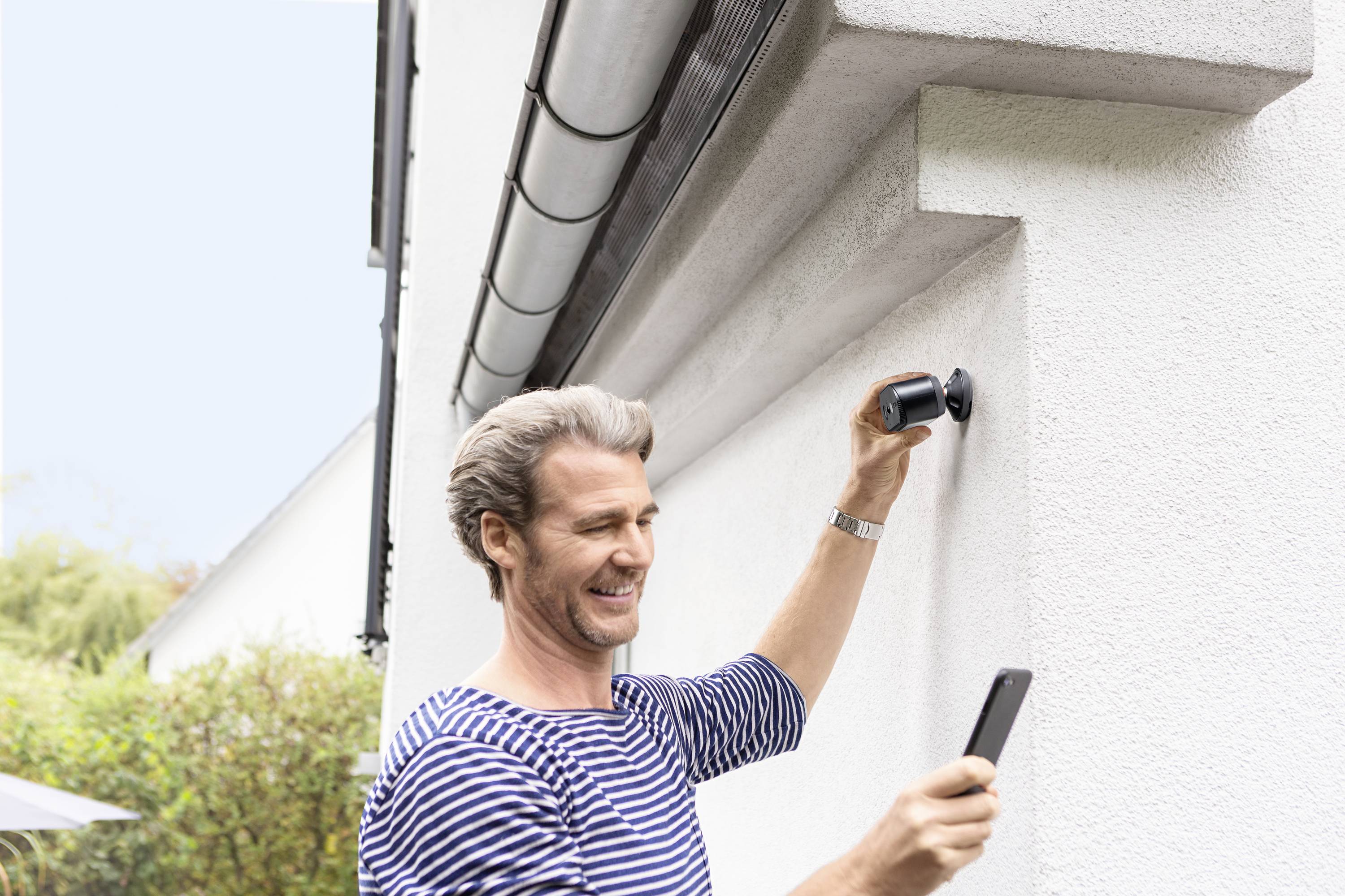 Een man bevestigt een camera aan een muur van een huis en kijkt naar zijn smartphone. De omgeving is groen en de lucht is helder.
