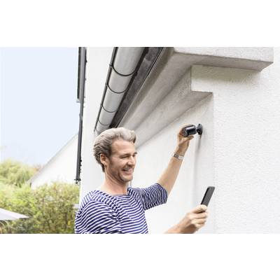 Een man bevestigt een camera aan een muur van een huis en kijkt naar zijn smartphone. De omgeving is groen en de lucht is helder.