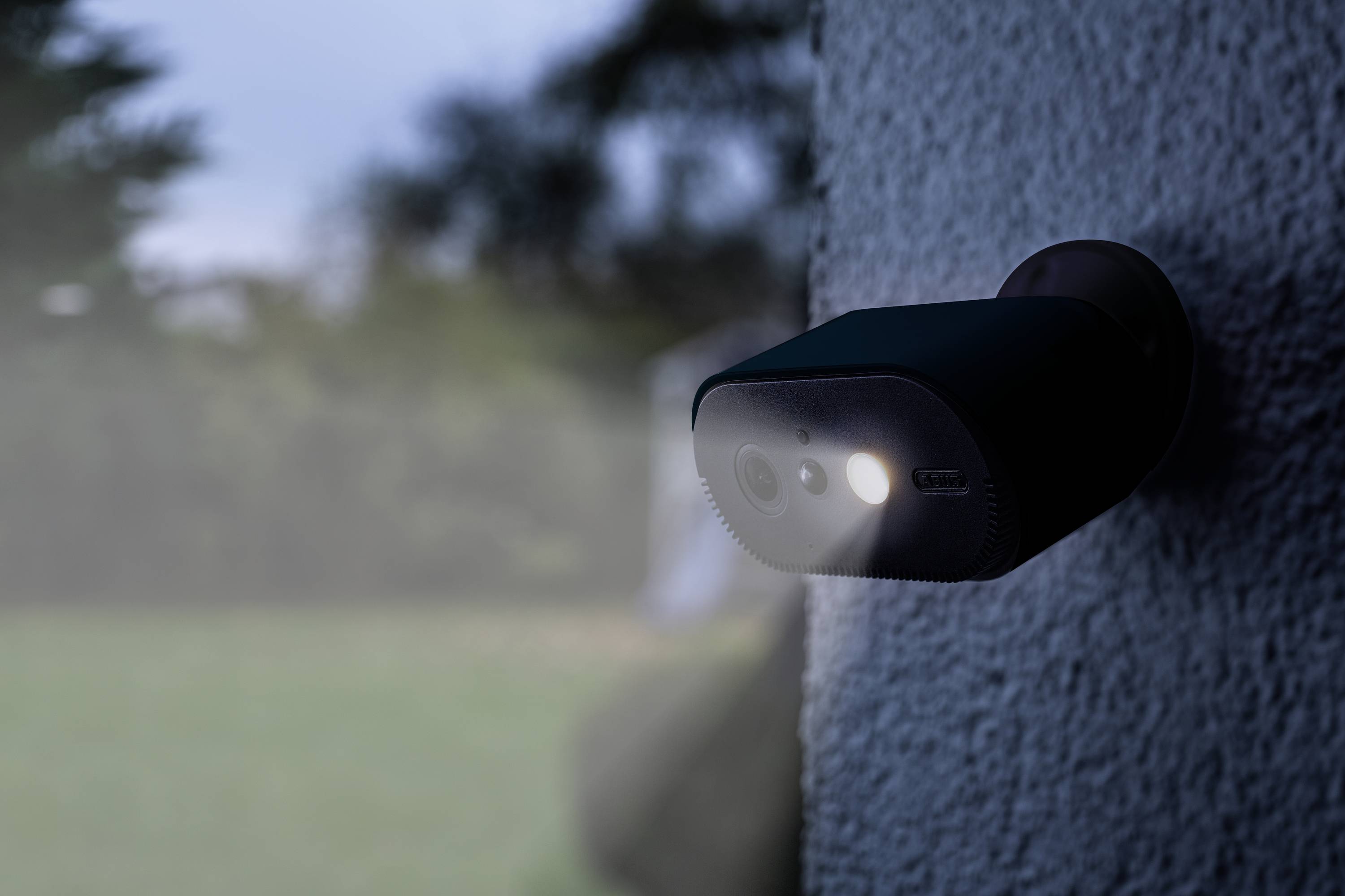 Buitencamera met nachtzicht en bewegingssensor, bevestigd aan een muur, in een tuin met een onscherpe achtergrond.