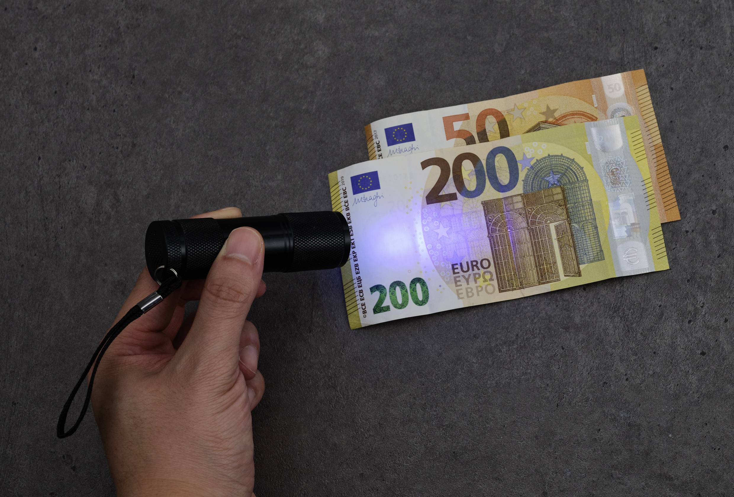 Een hand houdt een UV-zaklamp boven twee eurobiljetten (200 en 50 euro) om veiligheidskenmerken zichtbaar te maken.