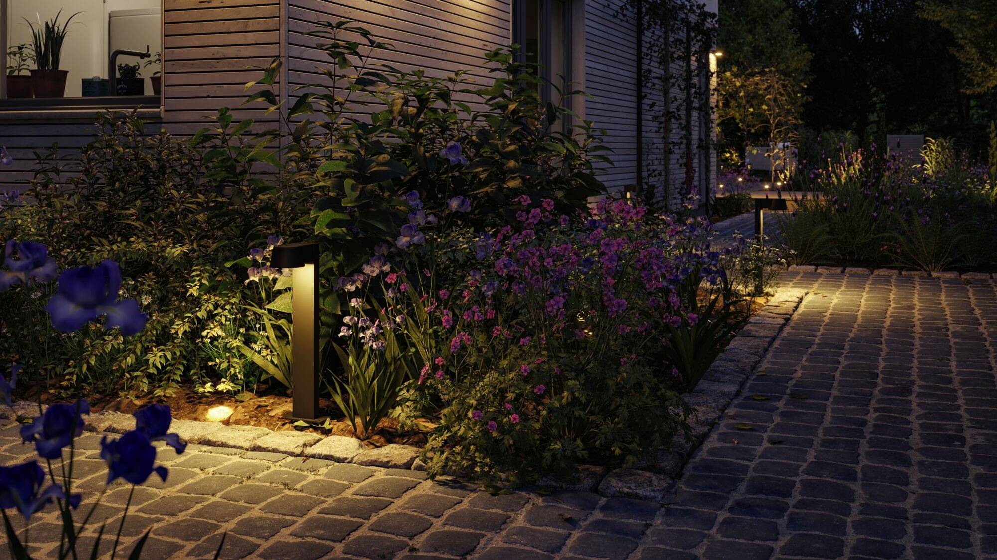 Tuinpad met kinderkopjes, omringd door bloeiende bloemen en moderne buitenverlichting, leidt naar een huis met houten planken.