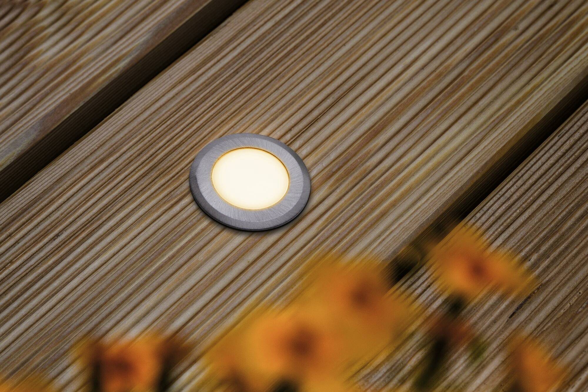 Een ronde LED-lamp is ingebouwd in een houten terras. Op de voorgrond zijn onscherpe oranje bloemen.