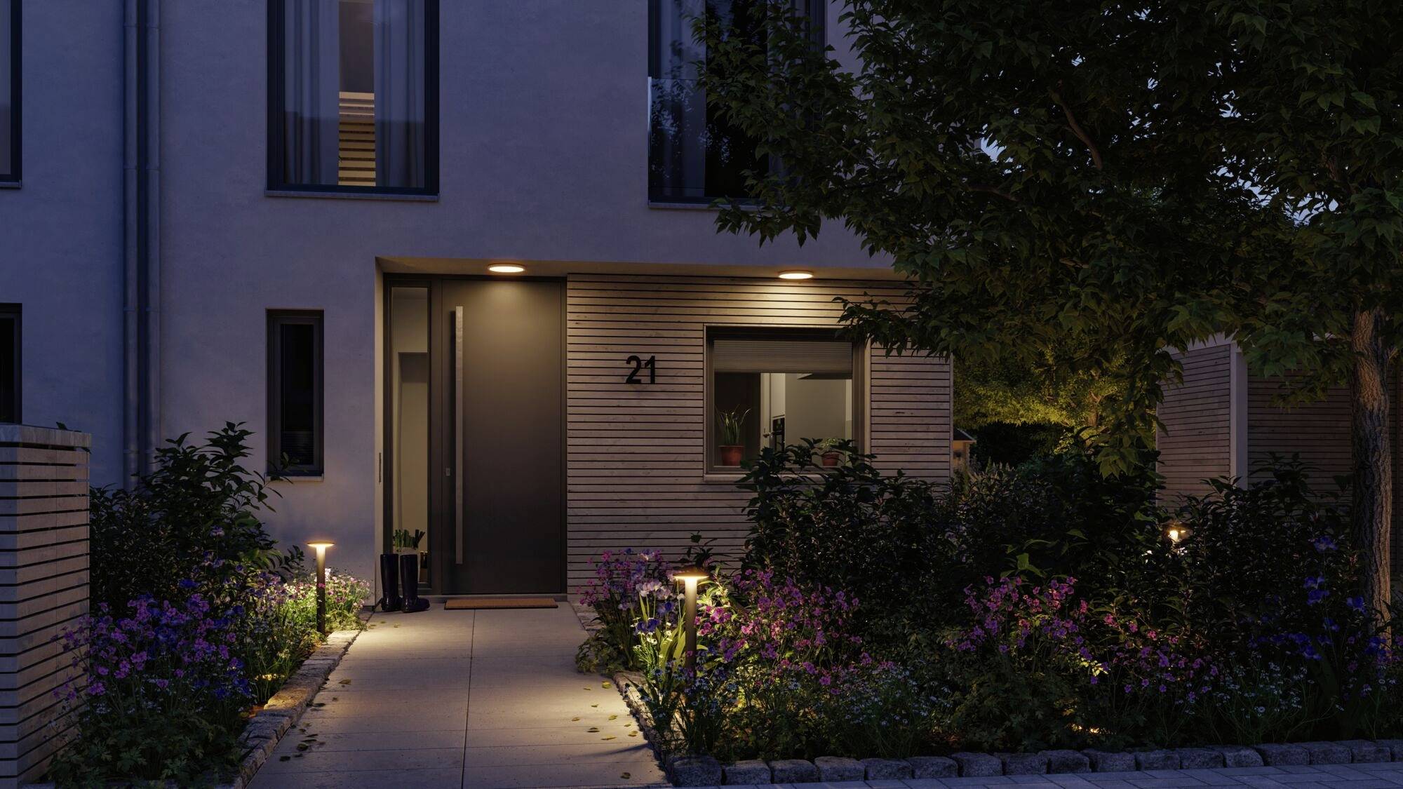 Moderne huisgevel 's avonds met verlichte ingang en keurig onderhouden tuin; huisnummer 21 zichtbaar.