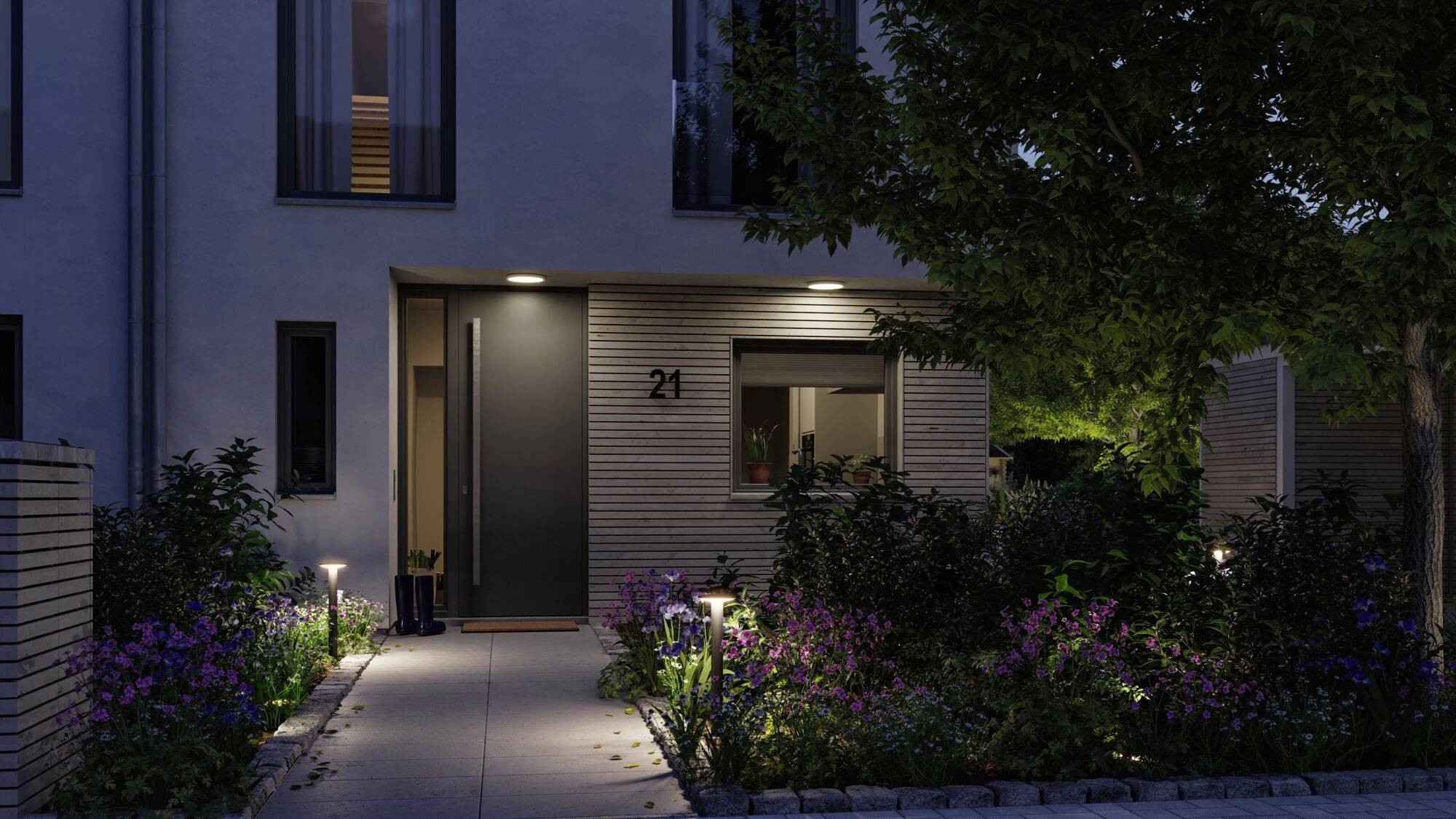 Ingang van een modern huis 's nachts, verlicht door verschillende tuinverlichting en omringd door planten. Huisnummer 21 zichtbaar links van de deur.