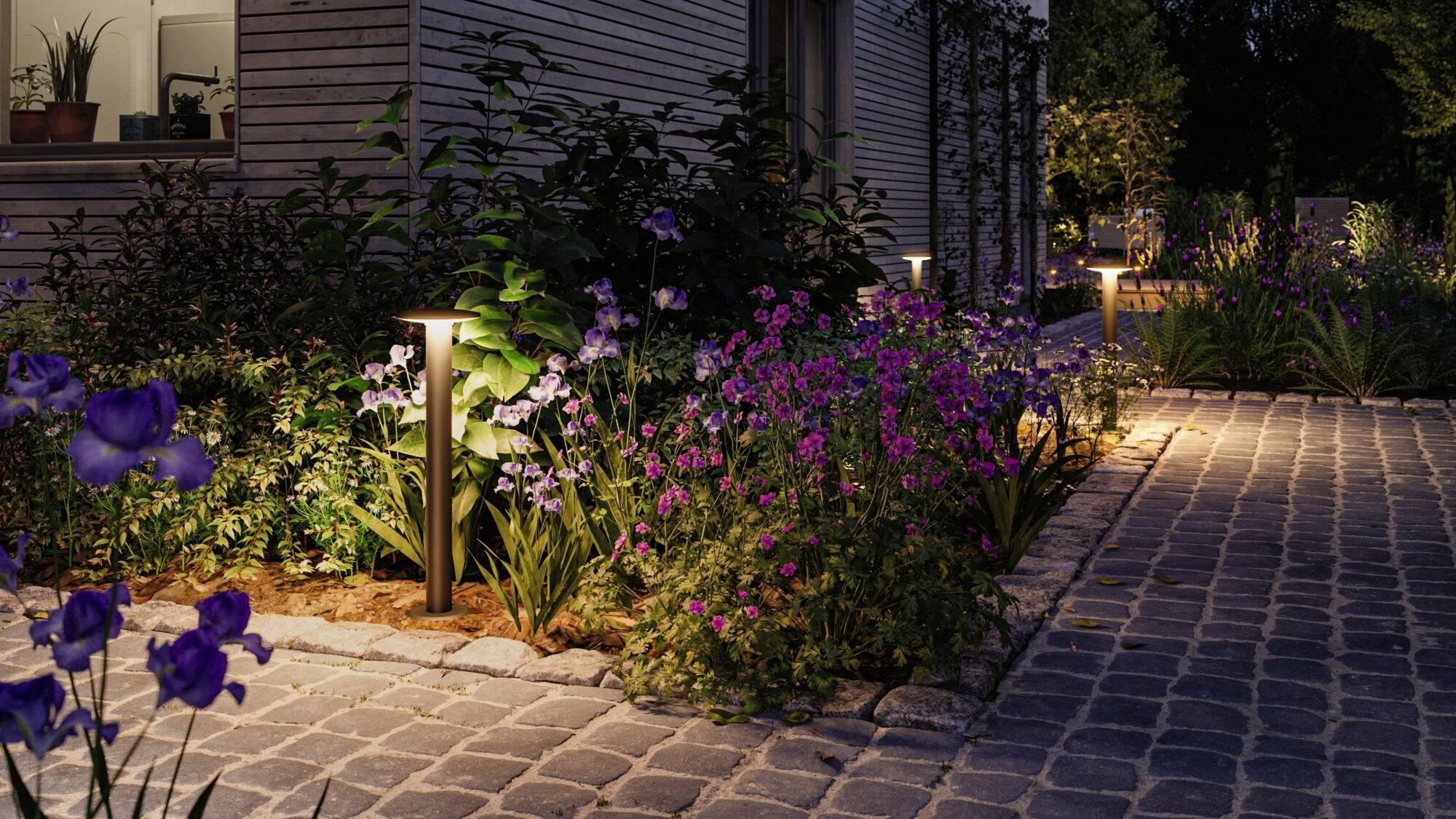 Verlichte tuinpad 's nachts, omzoomd door bloeiende, paarse en roze bloemen naast een houten huis.