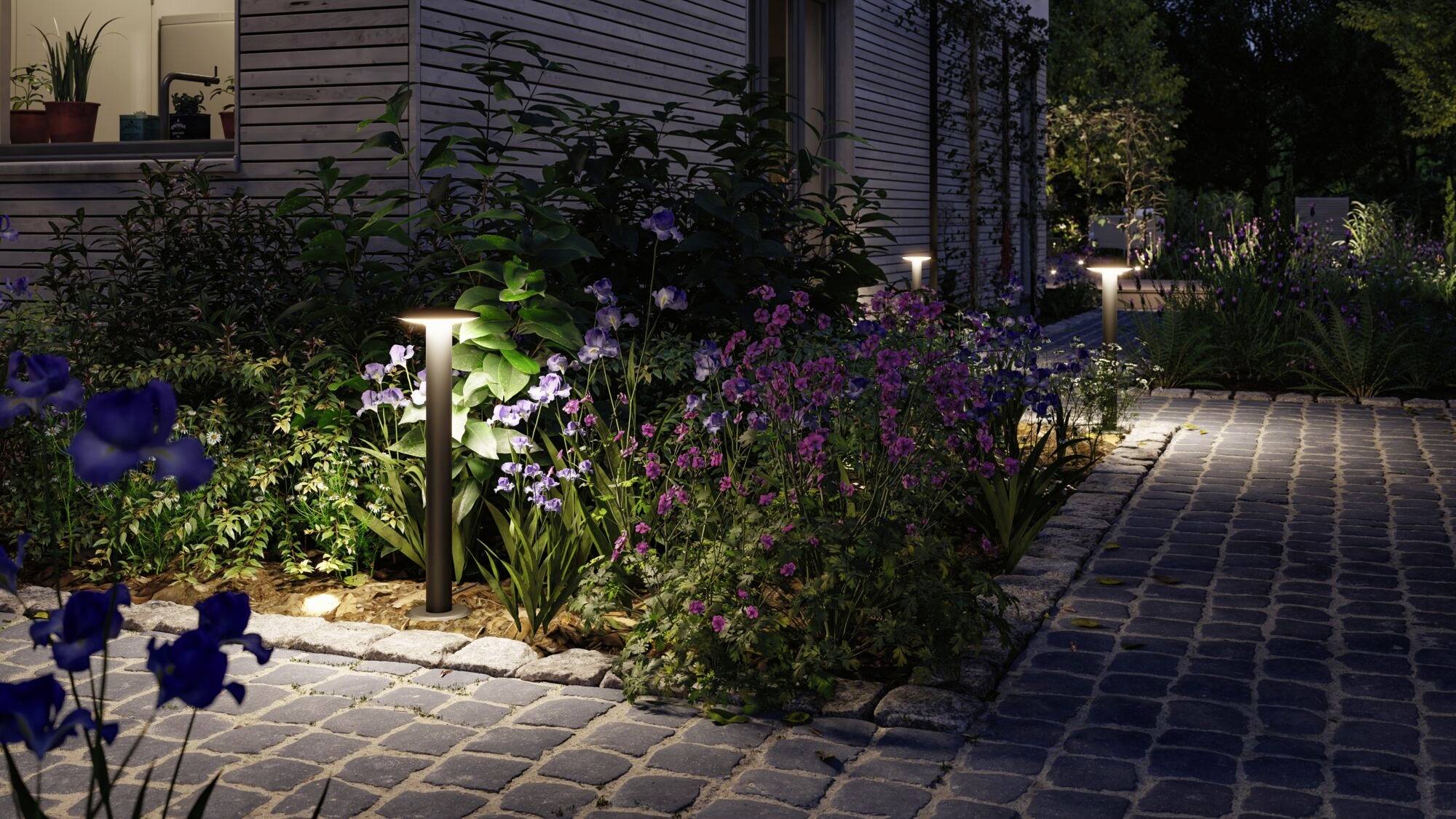 Een geplaveid tuinpad met verlichting 's nachts, omgeven door bloeiende bloemen en aangrenzende houten gebouwen.