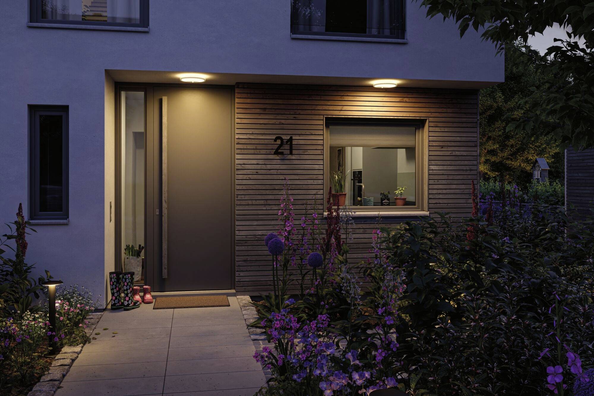 Ingang van een huis 's avonds, verlicht door buitenlampen. Bloemperken flankeren het pad naar de deur. Ramen met planten aan de binnenkant.