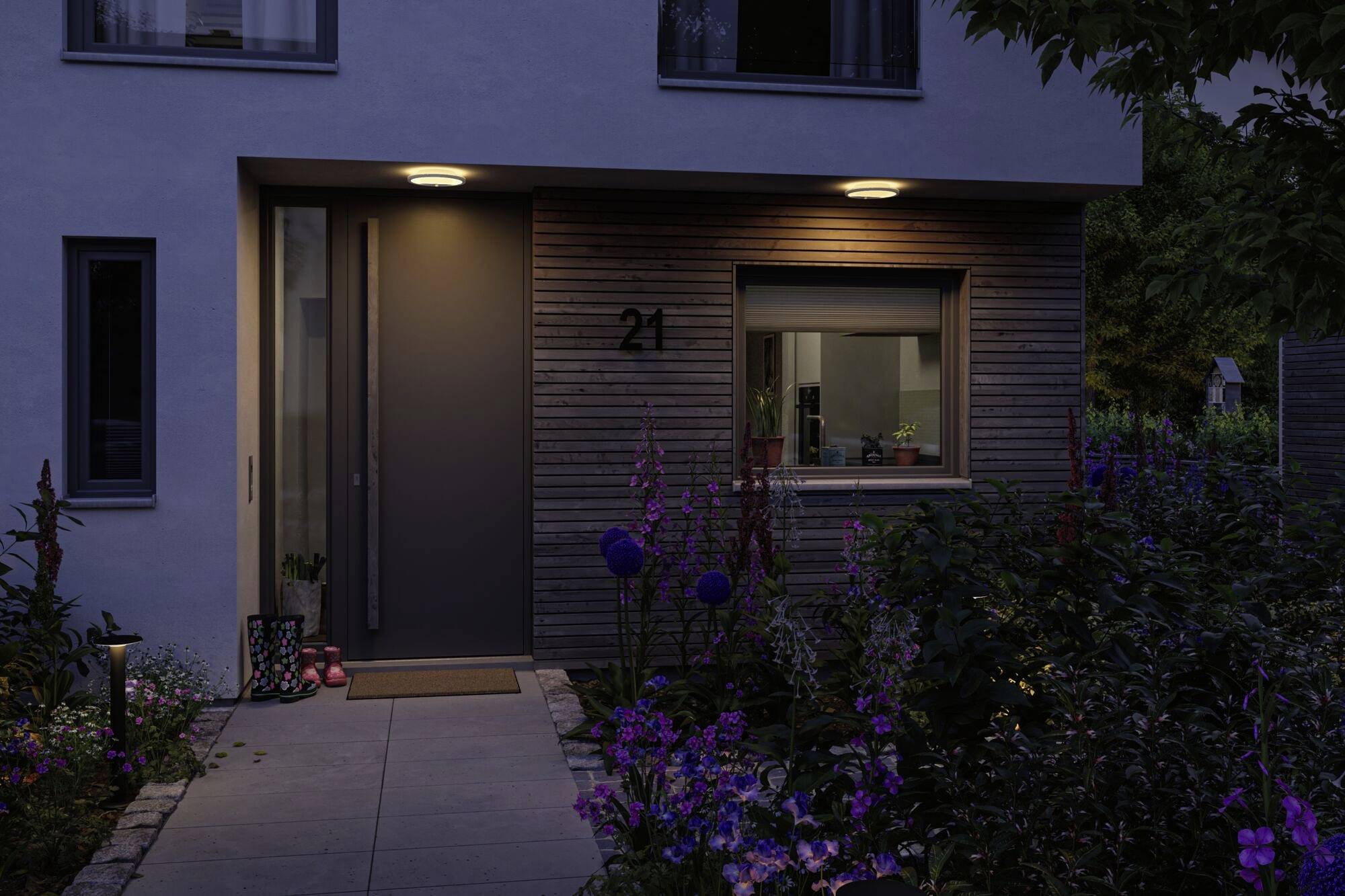 Een verlichte entree van een huis 's avonds, met moderne houten accenten en een verzorgde tuin vol kleurrijke bloemen op de voorgrond.