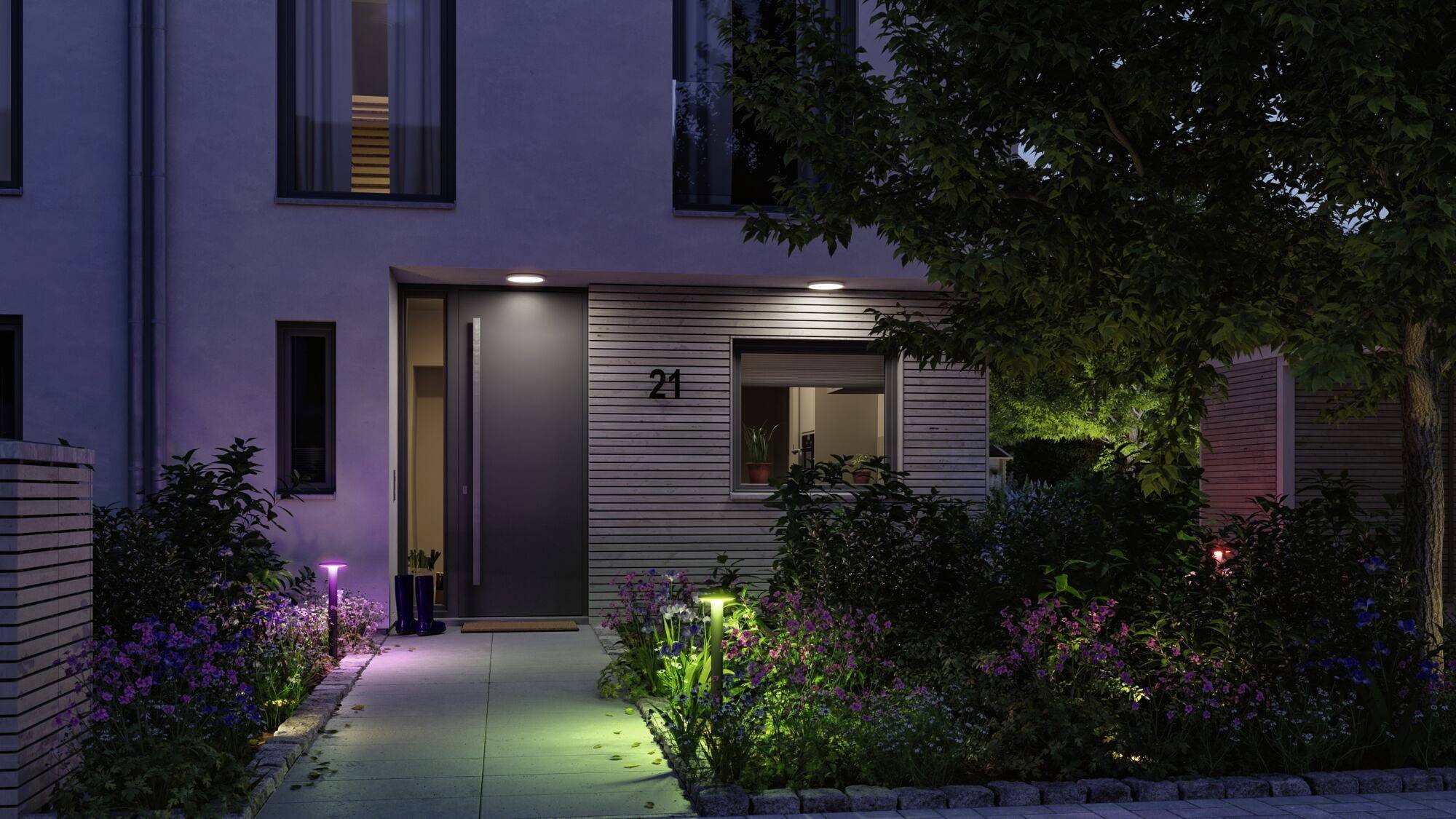 Woonhuis 's avonds met verlichte ingang. Tuinplanten flankeren het pad naar de deur. Huisnummer 21 is zichtbaar.
