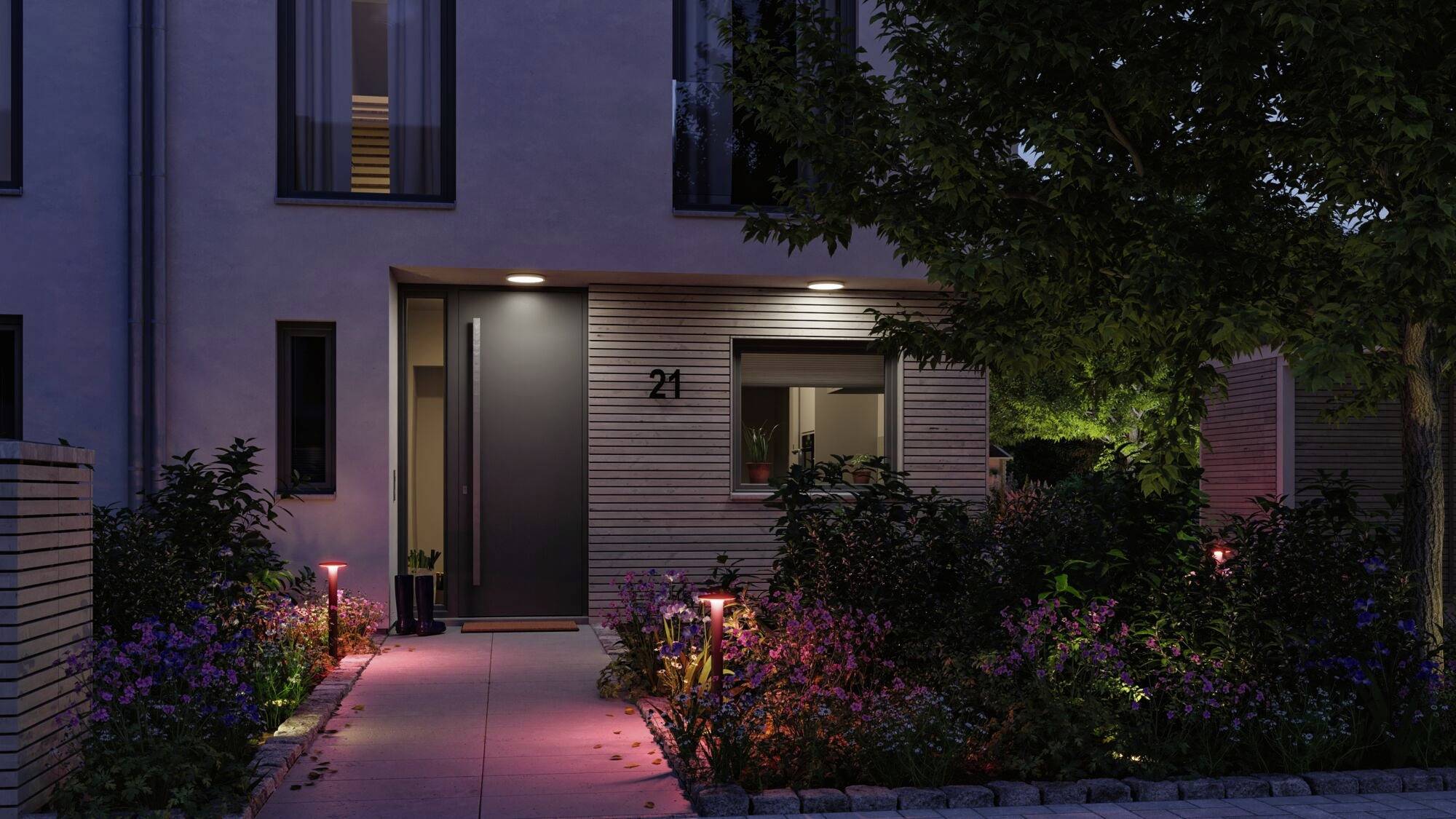Een modern gebouw 's nachts met een verlichte ingang en huisnummer 21, omgeven door planten en subtiele tuinverlichting.