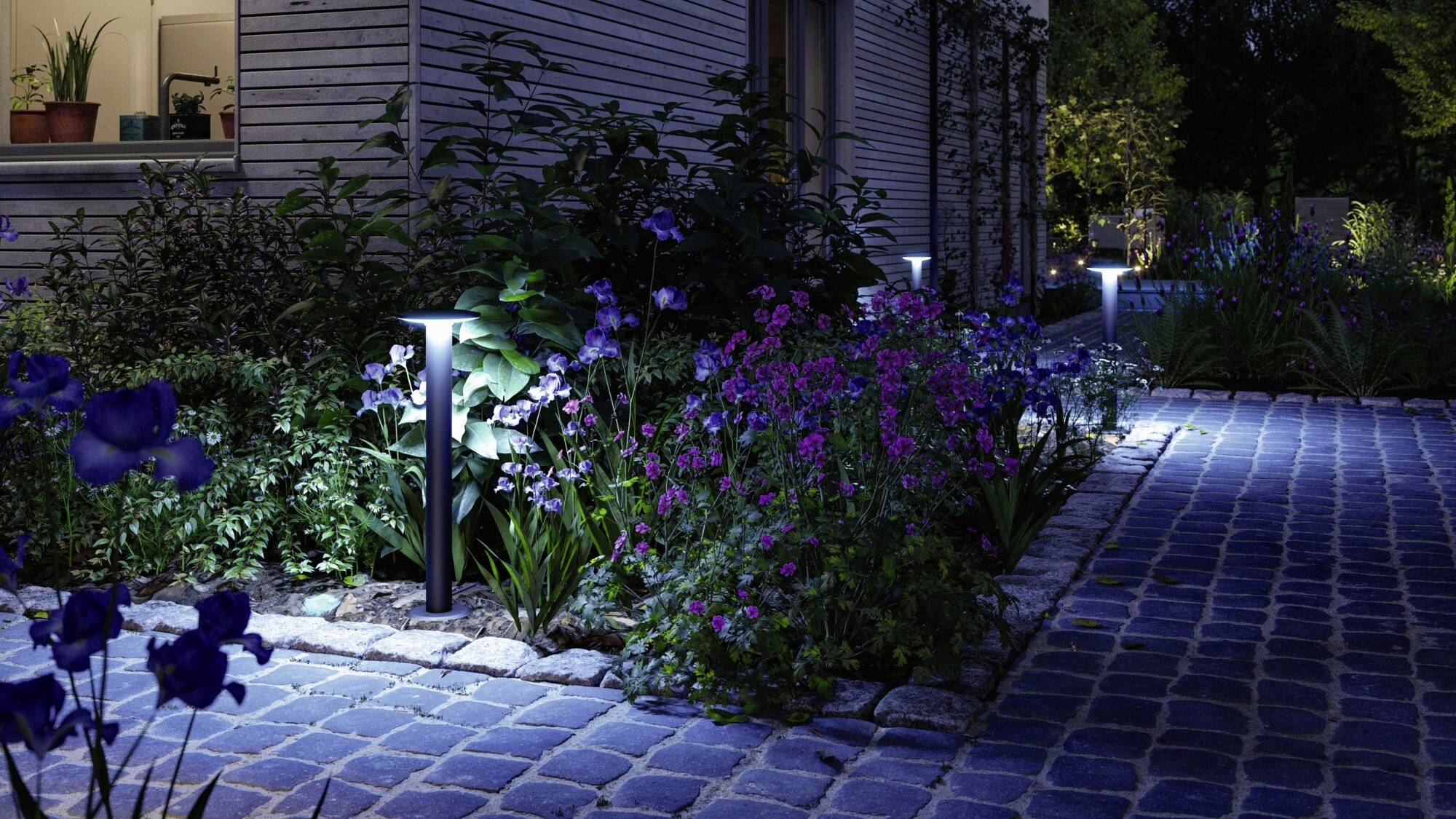 Een verlicht tuinpad van steenkeien leidt langs bloeiende planten die zacht worden verlicht in de avondschemering.