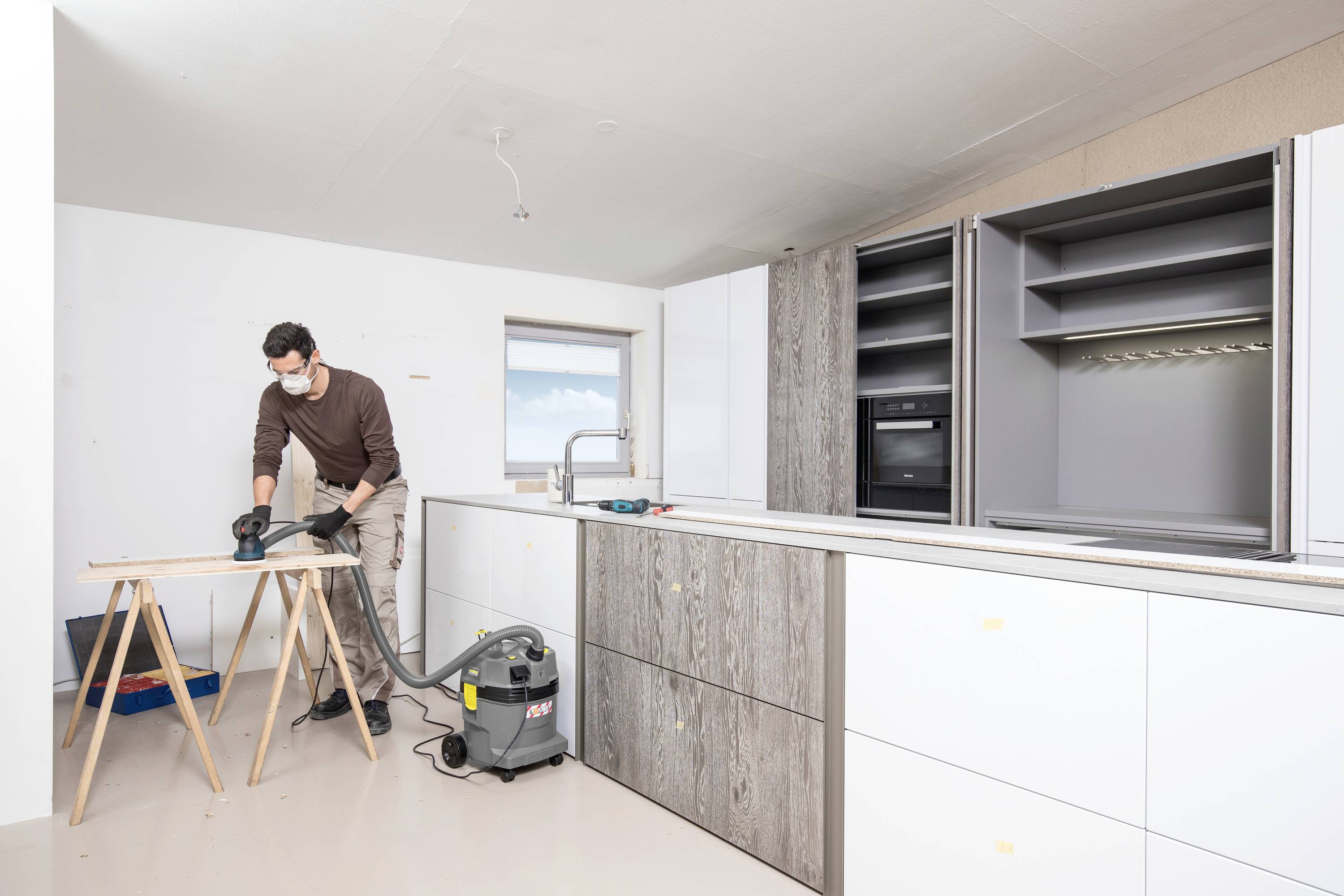 Een man werkt aan een houten snijplank in een lichte keuken. Hij draagt een beschermingsmasker en handschoenen. Naast hem staat een stofzuiger.