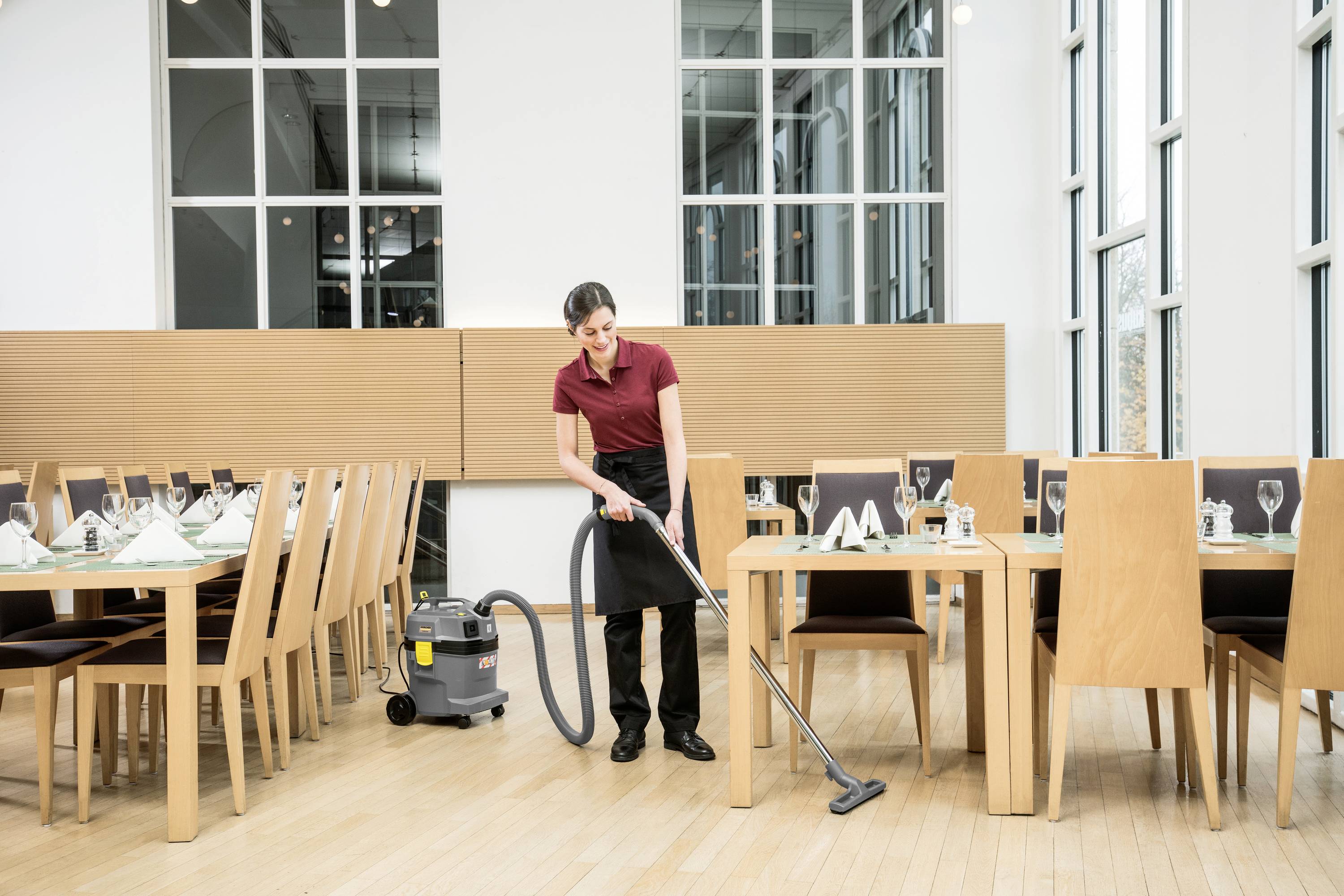 Een persoon zuigt de vloer in een leeg restaurant met een tafelstofzuiger. Tafels zijn gedekt, grote ramen op de achtergrond.