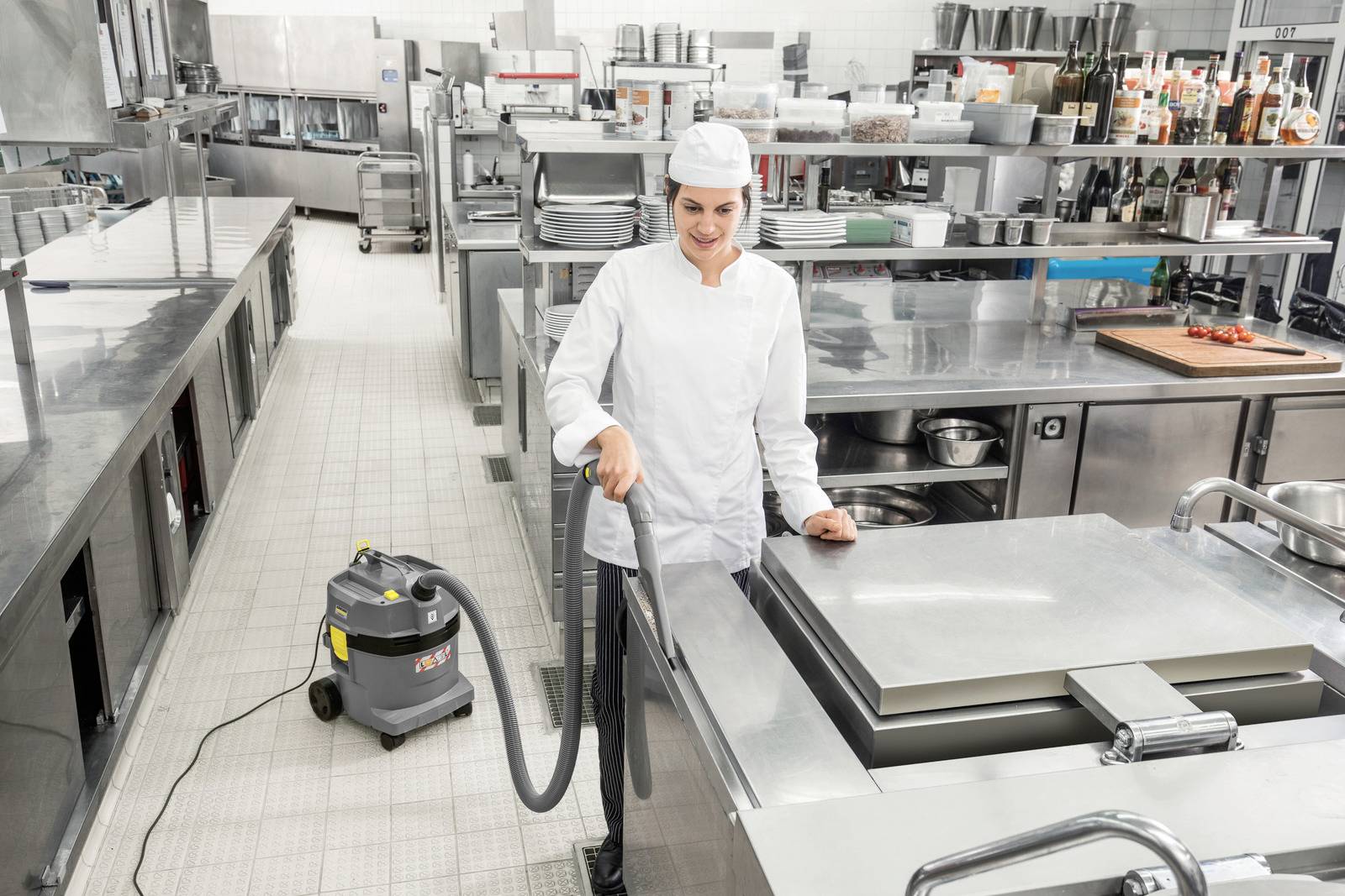 'Een chef in een commerciële keuken gebruikt een stofzuiger om roestvrijstalen oppervlakken schoon te maken, omringd door industriële apparatuur en kookgerei.'