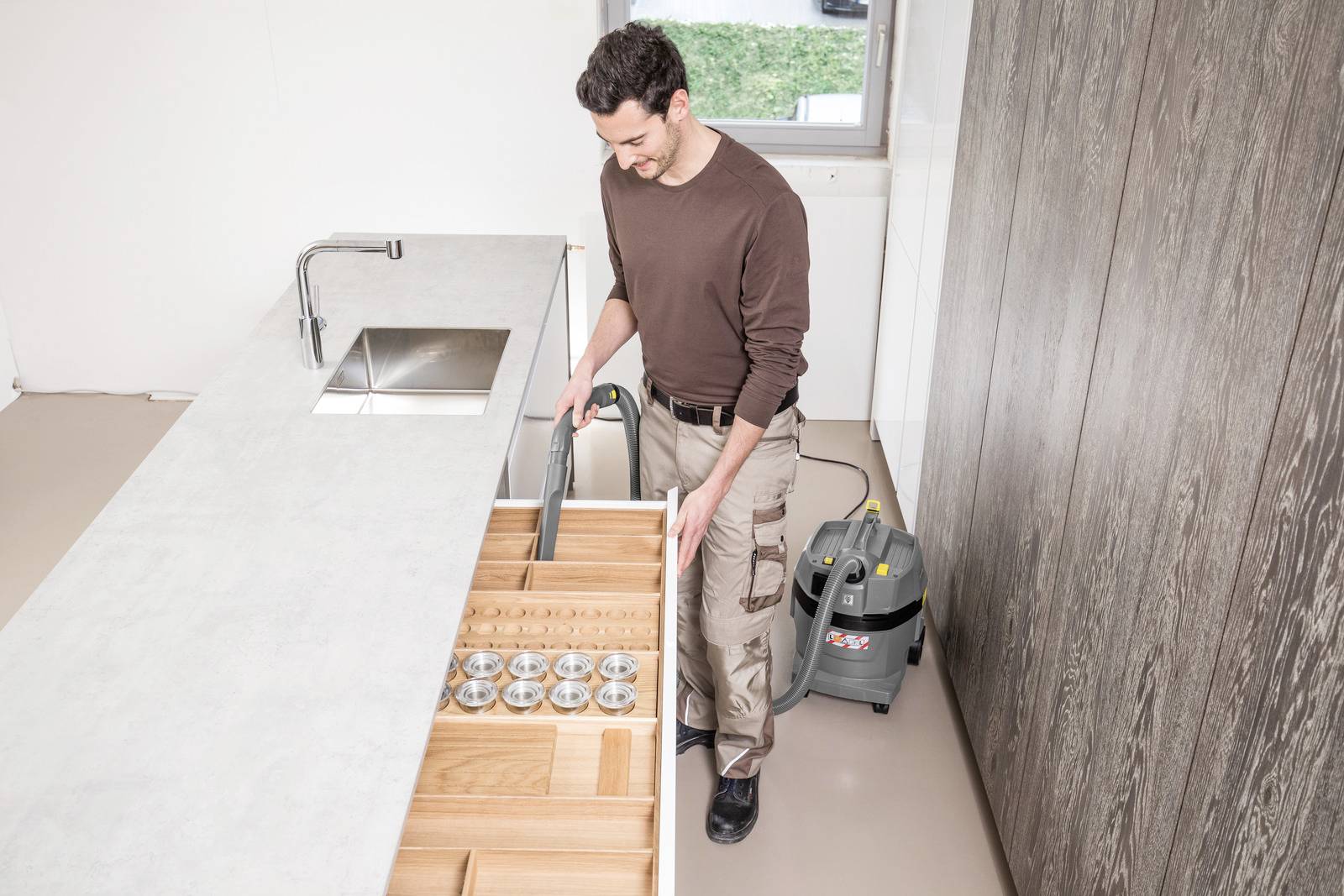 Een man die een stofzuiger gebruikt om een houten lade schoon te maken in een moderne keukenomgeving; een stofzuiger staat op de vloer gepositioneerd.