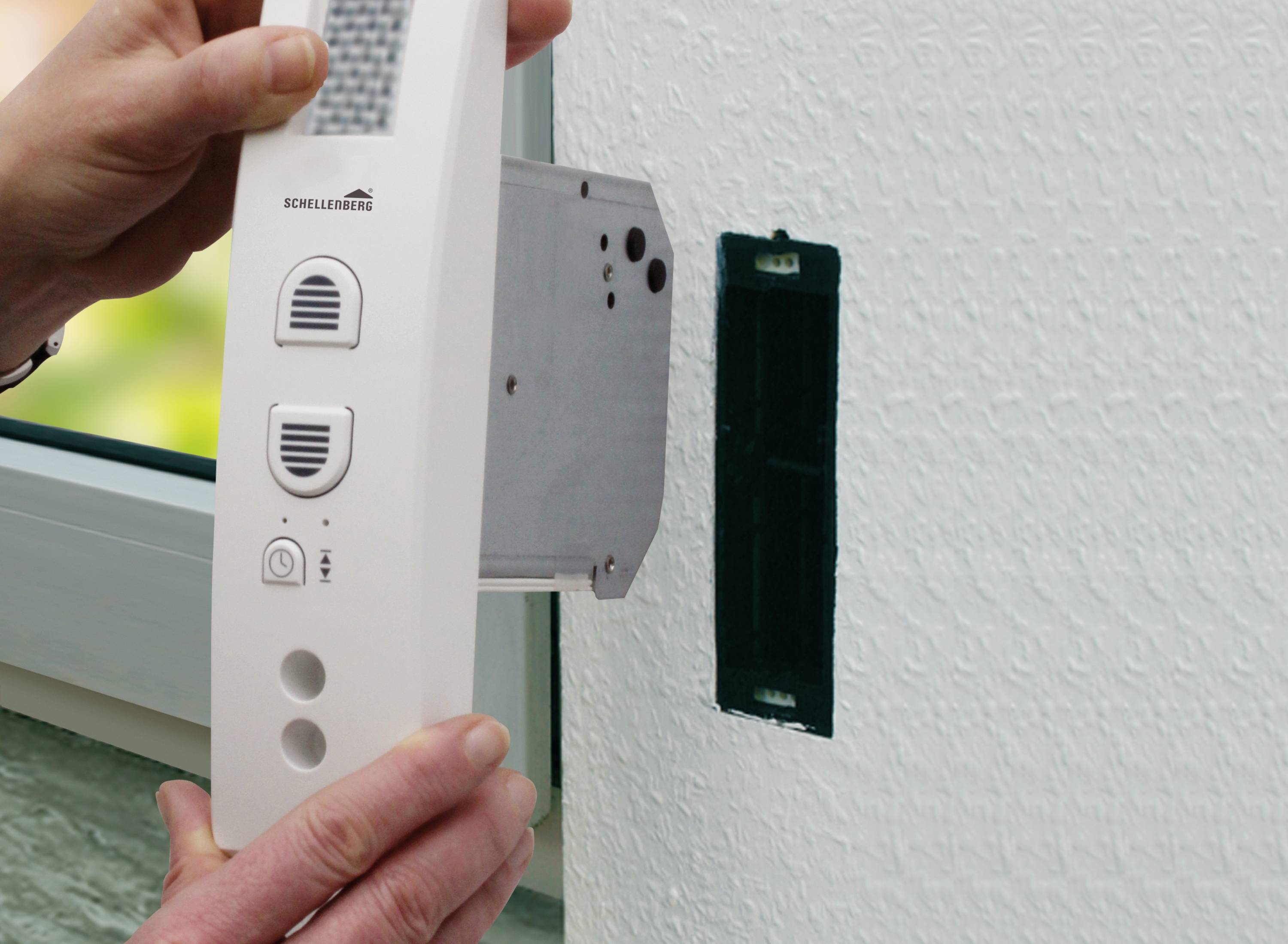 Een persoon installeert een witte intercominstallatie aan de muur naast een raam. De intercompost heeft knoppen en een luidsprekerooster.