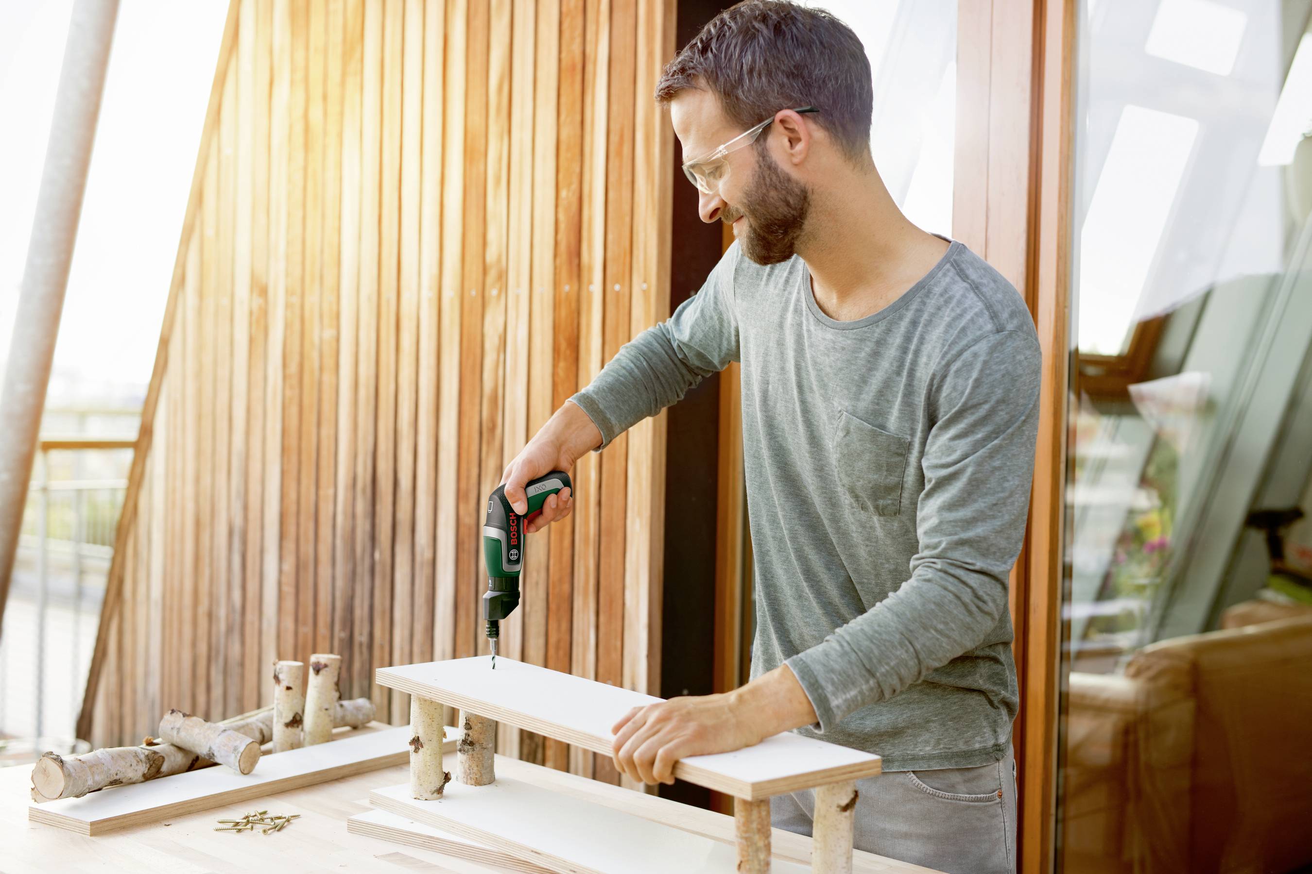 Man met een bril boort gaten in een houten plank met een elektrische boormachine, terwijl hij werkt aan een doe-het-zelf project op een terras.