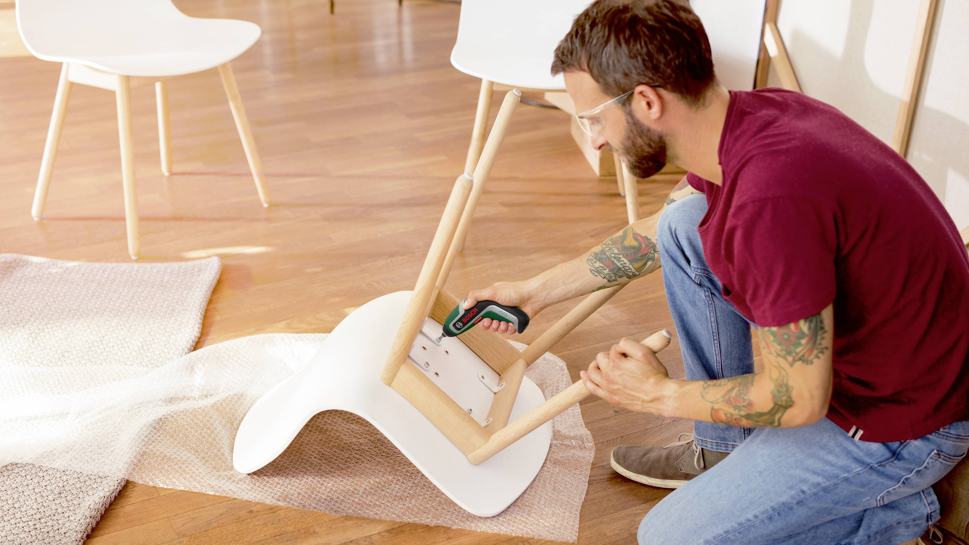 Een man monteert een witte stoel met een accuboormachine in een goed verlichte ruimte met een houten vloer en andere stoelen op de achtergrond.