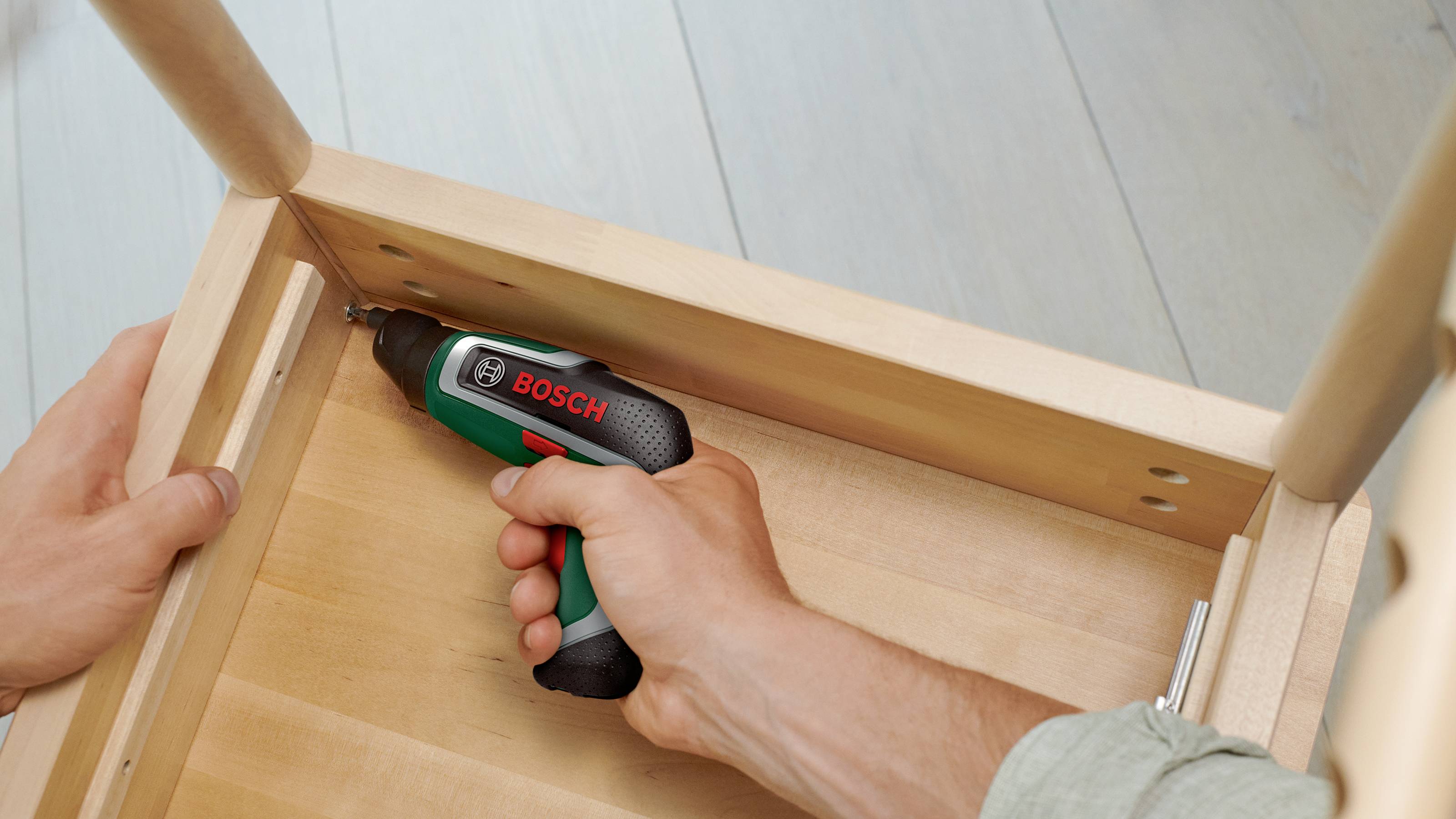 Een persoon monteert een houten tafel met een Bosch accuschroefmachine door een schroef aan te draaien. De tafel bevindt zich in een ruimte met een houten vloer.