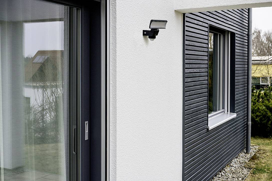 Een modern huis met witte en grijze muren; een bewegingsmelder is naast een raam en een glazen deur aangebracht.