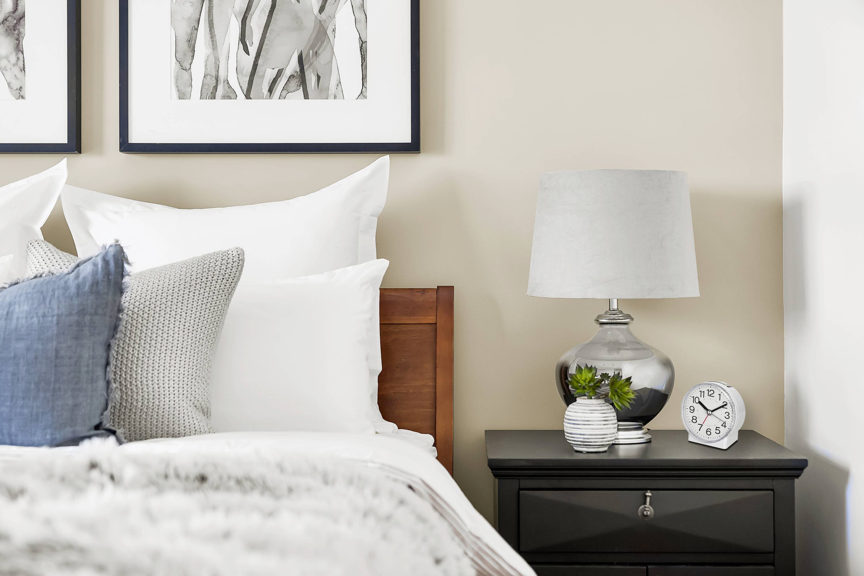 Een slaapkamer met een bed, versierd met witte en blauwe kussens. Naast het bed staat een nachtkastje met een lamp, een plant en een wekker.