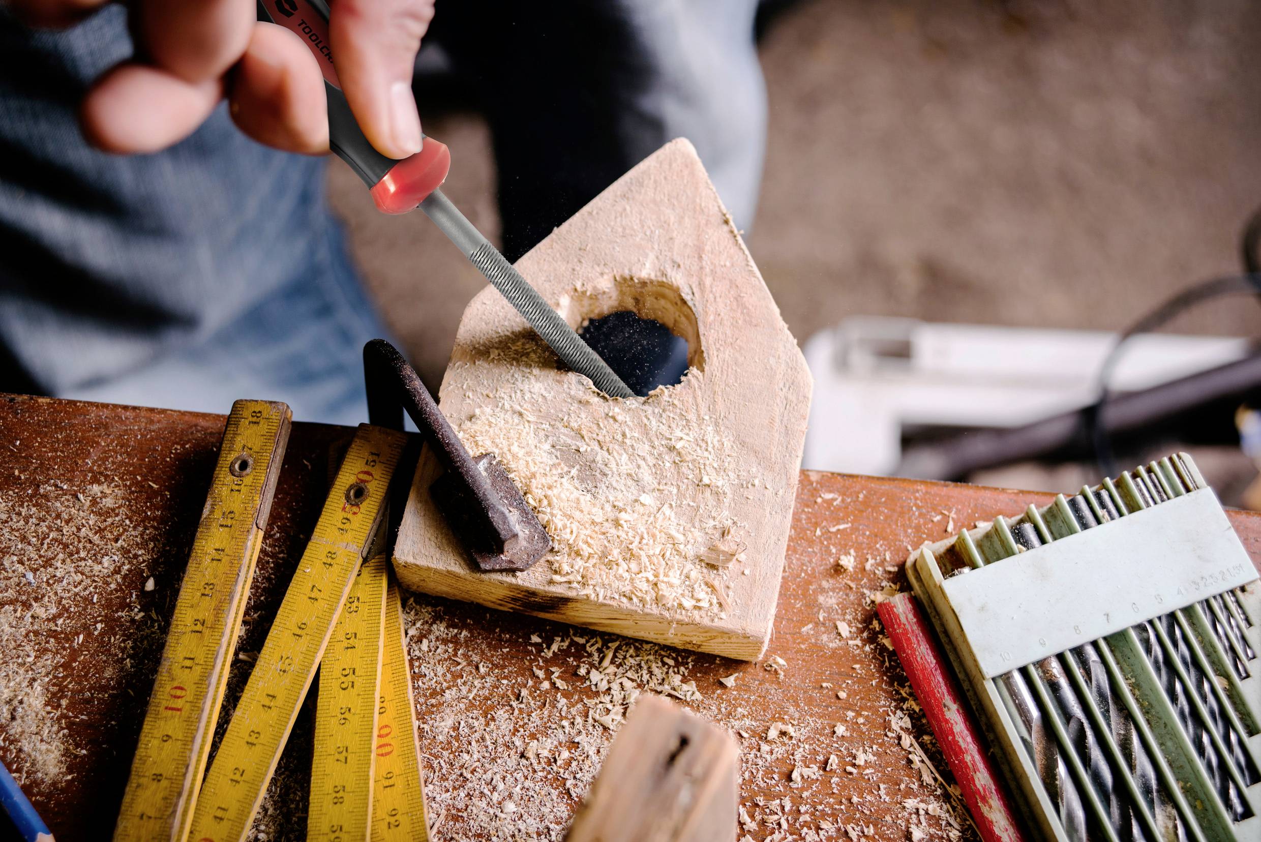 Een persoon vijlt een gat in een stuk hout. Omringd door een meetlint, houtkrullen en een notitieblok. Focus op handmatig werk.