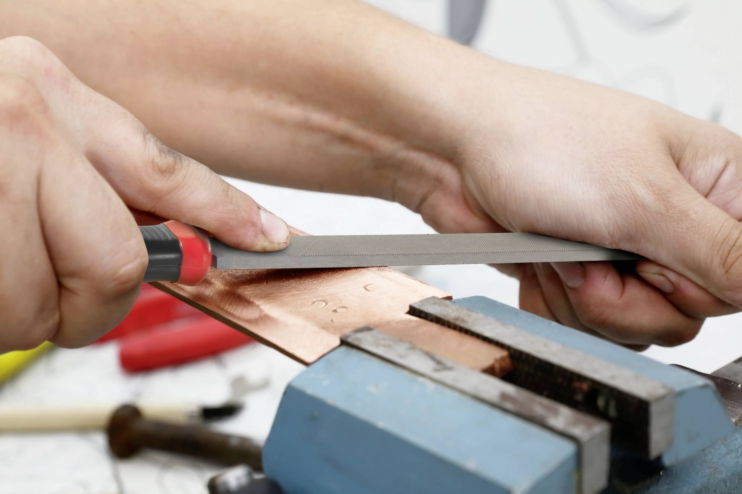 Een persoon vijlt een stuk koper dat in een bankschroef is geklemd, in een werkplaats.