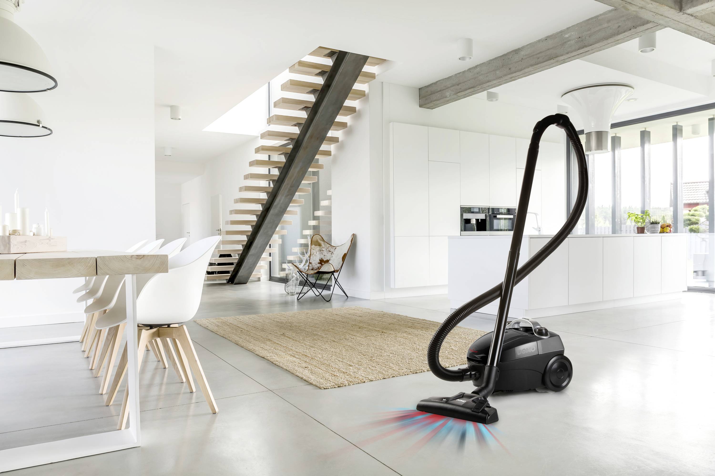 Een stofzuiger staat in een modern woonkamer met witte meubels en een grote trap, klaar om de vloer schoon te maken.