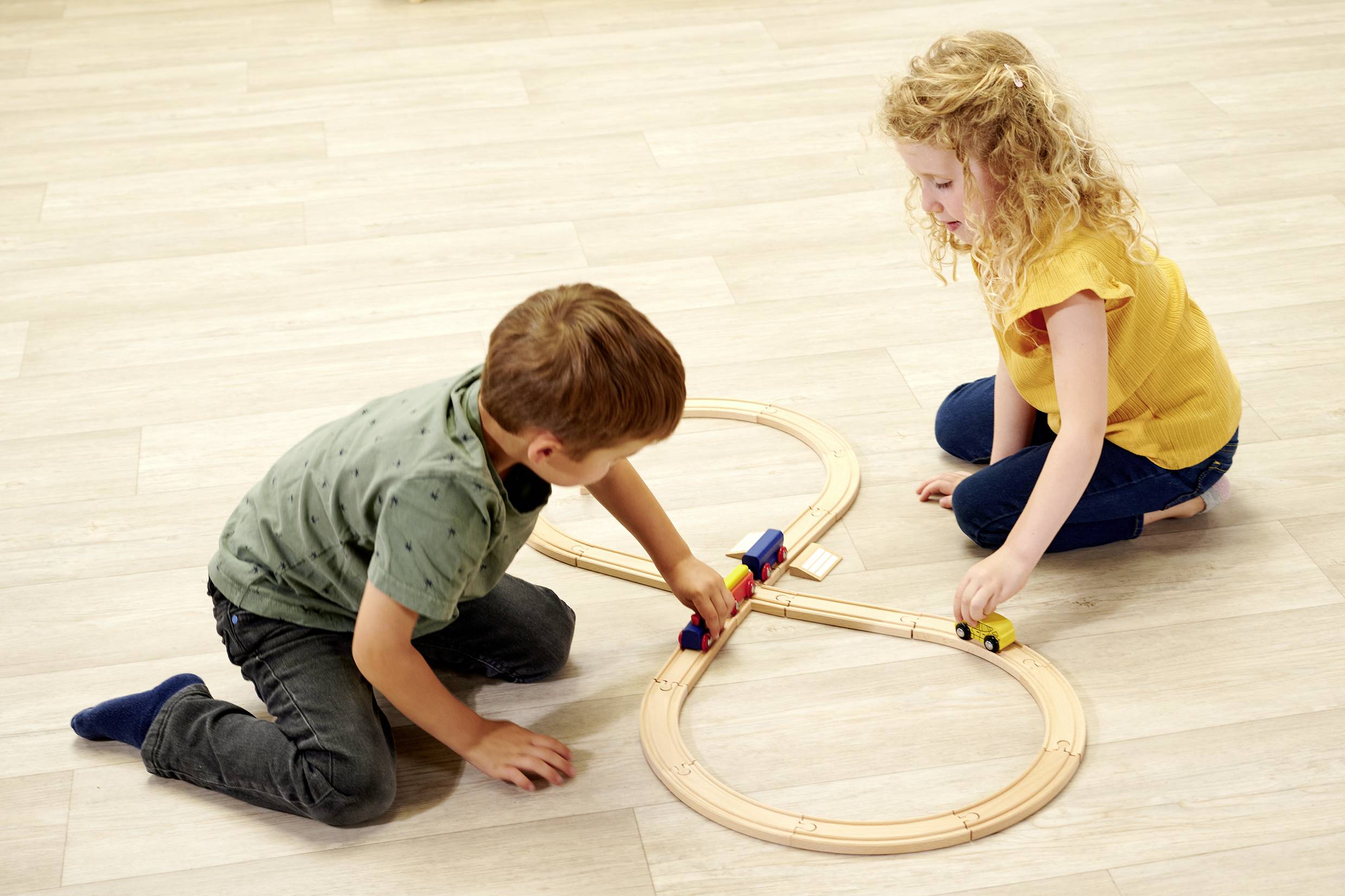 Twee kinderen spelen op een houten vloer met een houten baan in de vorm van een acht, een jongen bestuurt een blauwe trein, een meisje een gele.