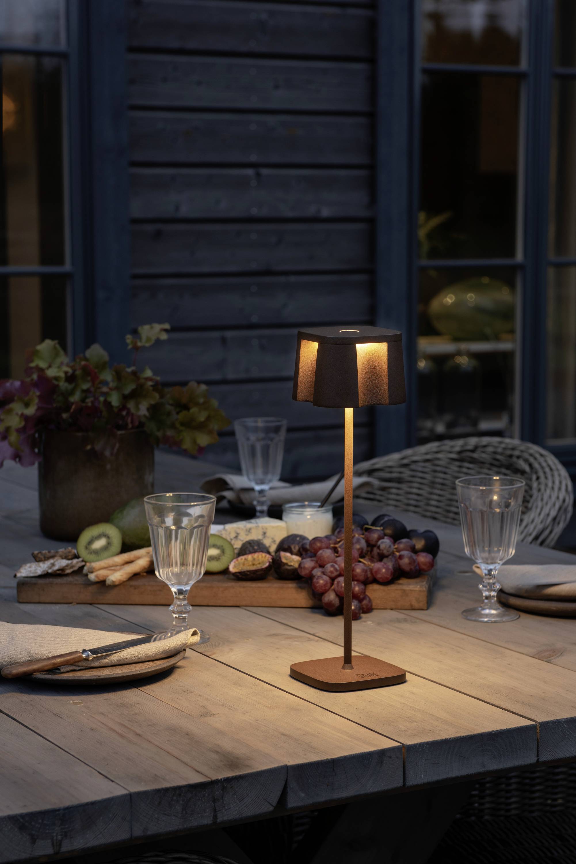 Een tafellamp verlicht een gedekte houten tafel buiten met een fruitschaal, kiwi, druiven en lege glazen. Gedempte avondverlichting.