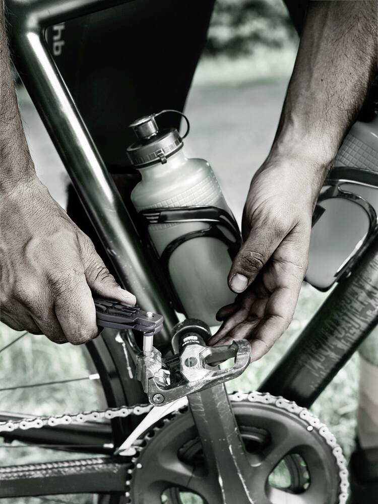 Persoon repareert fietsversnelling met gereedschap; waterbidon bevestigd aan het frame; focus op handen en pedaal.